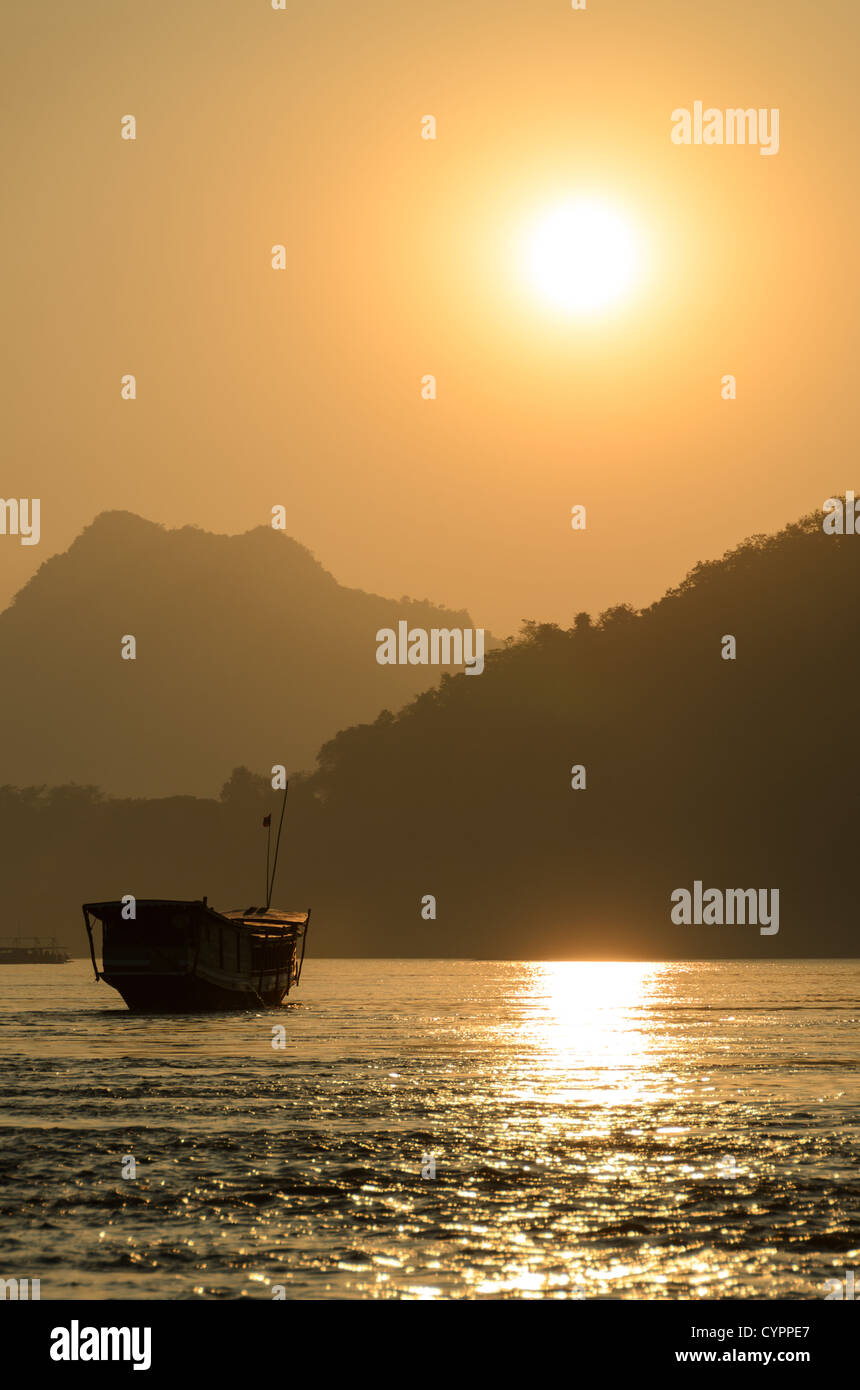 Barca tradizionale del fiume Mekong Luang Prabang Laos // LUANG PRABANG, Laos - il fiume Mekong scorre oltre questa città patrimonio dell'umanità dell'UNESCO mentre le tradizionali barche in legno attraversano il corso d'acqua in lontananza. La luce dorata filtra attraverso la foschia atmosferica durante il tramonto, creando sagome delle montagne circostanti. Luang Prabang si trova alla confluenza dei fiumi Mekong e Nam Khan nel Laos settentrionale. La città è stata la capitale reale del Regno del Laos fino al 1975 e rimane un importante centro culturale. Il trasporto fluviale tradizionale continua a svolgere un ruolo fondamentale nel commercio locale e quotidiano Foto Stock