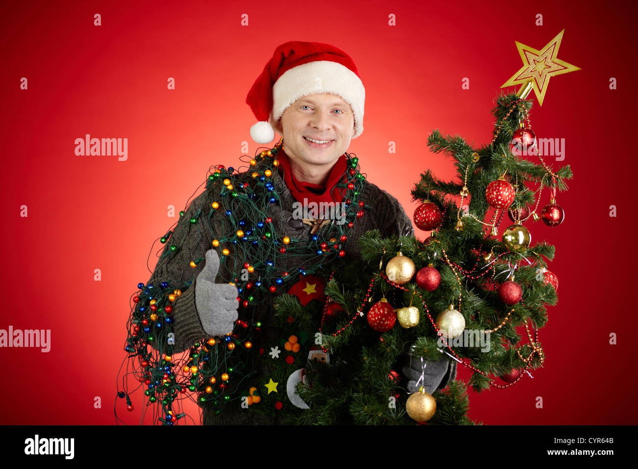 Ritratto di uomo felice in Santa cappuccio decorate albero di Natale che mostra il pollice in alto Foto Stock