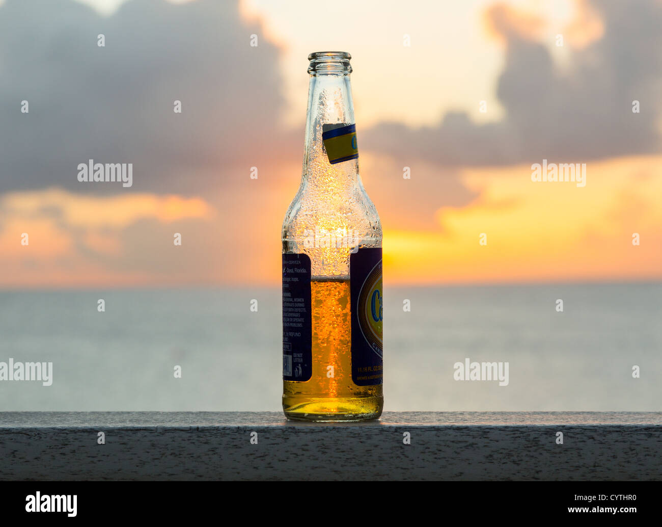 Di ghiaccio freddo Carib bottiglia di birra sul tavolo di fronte tramonto sull'oceano Foto Stock