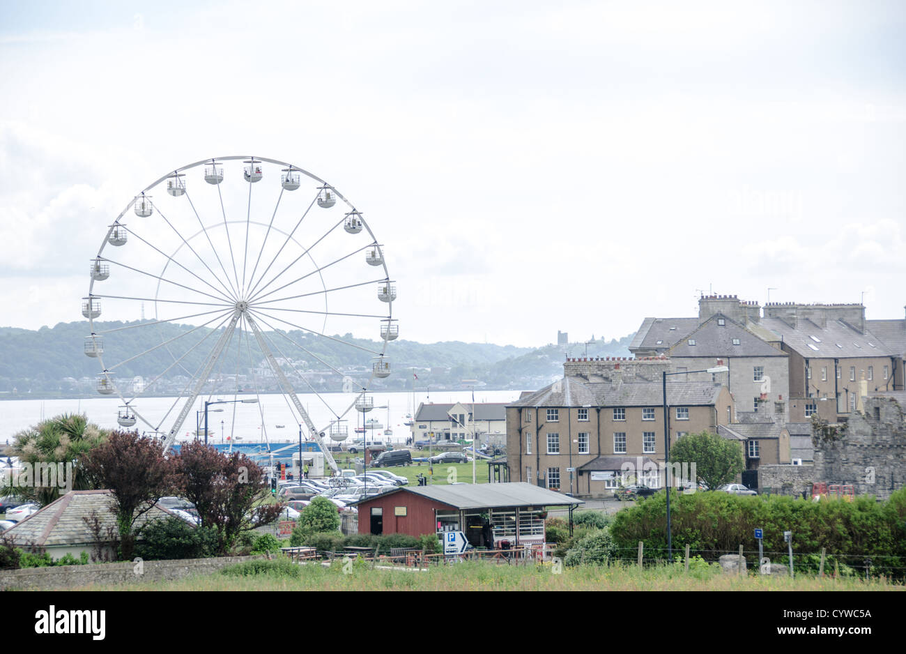 BEAUMARIS, Galles — Una ruota panoramica si erge in modo prominente tra i tetti di Beaumaris sull'isola di Anglesey. La città costiera si trova lungo lo stretto di Menai, il corpo d'acqua che separa Anglesey dal Galles continentale. Beaumaris, situata sulla costa sud-orientale dell'isola, è conosciuta per il suo castello del XIII secolo costruito da Edoardo i durante la sua conquista del Galles. La città è una destinazione popolare su Anglesey, la più grande isola del Galles. Lo stretto di Menai, visibile oltre la città, è una stretta via d'acqua che ha storicamente fornito importanza strategica alla regione. Foto Stock