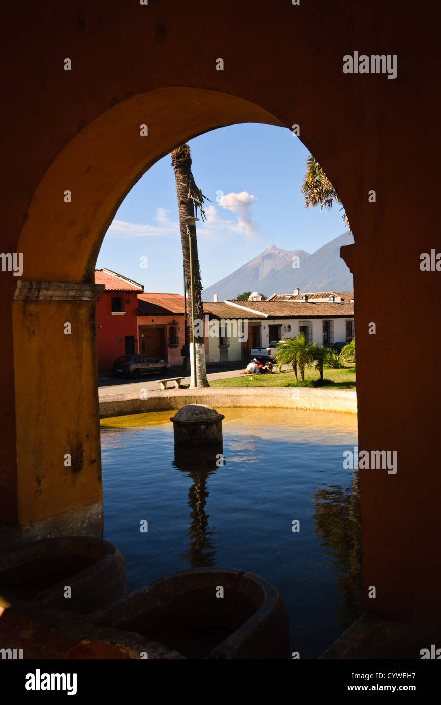 Fonte d'acqua pubblica Volcano Vista Antigua Guatemala // ANTIGUA, Guatemala — Una fonte d'acqua pubblica o un'area di lavaggio si trova lungo una strada acciottolata ad Antigua, Guatemala, con un vulcano visibile sullo sfondo che emette un pennacchio di fumo. La struttura in pietra, probabilmente una delle storiche fontane pubbliche della città o "pilas" che un tempo serviva come fonti d'acqua della comunità, riflette l'infrastruttura coloniale dell'ex capitale. Antigua, patrimonio dell'umanità dell'UNESCO dal 1979, è conosciuta per la sua architettura coloniale spagnola ben conservata ed è circondata da tre vulcani, tra cui Volcán de Agua Foto Stock