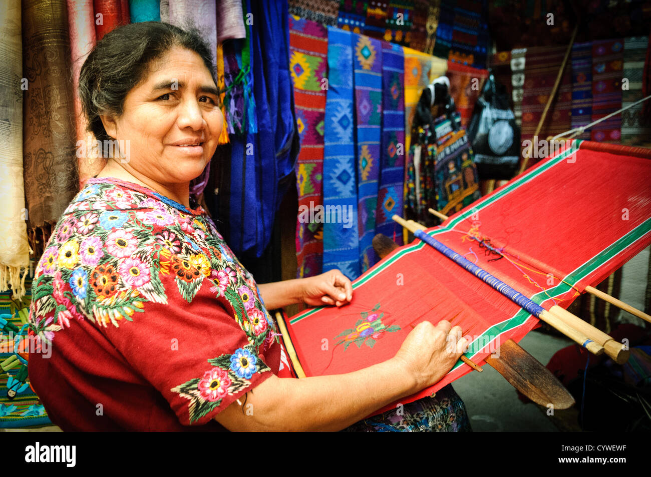 ANTIGUA GUATEMALA, Guatemala - Una donna Maya dimostra la tradizionale tessitura del telaio dello schienale, creando tessuti vivaci. Le sue mani manipolano abilmente fili colorati, producendo intricati motivi che riflettono tecniche secolari e simbolismo culturale, mentre huipiles finiti e altri tessuti sono appesi sullo sfondo. Foto Stock