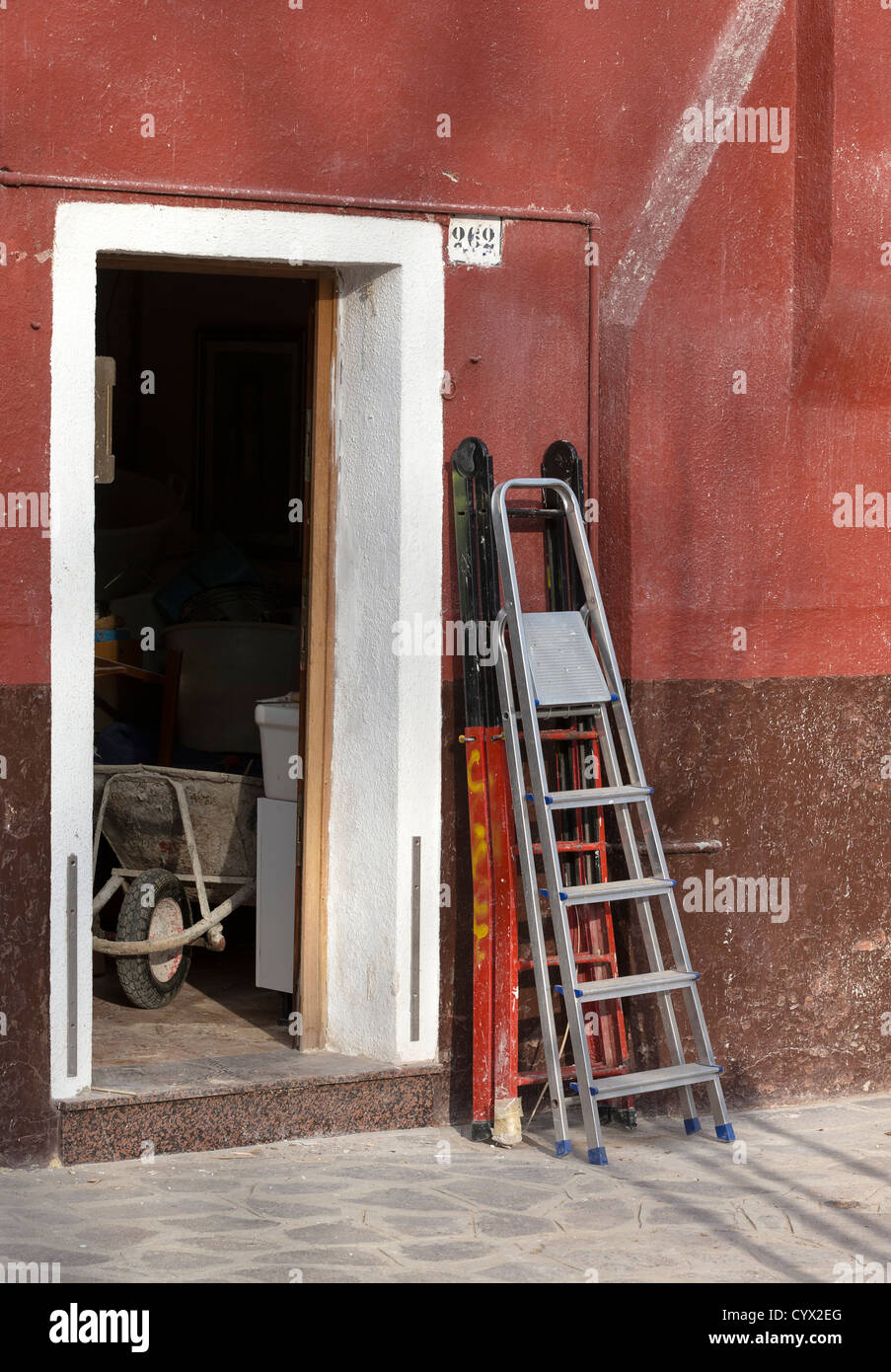 Scale a gradini appoggiata contro una parete a Burano, Venezia, con una carriola visibili all'interno della porta che indica i costruttori al lavoro Foto Stock