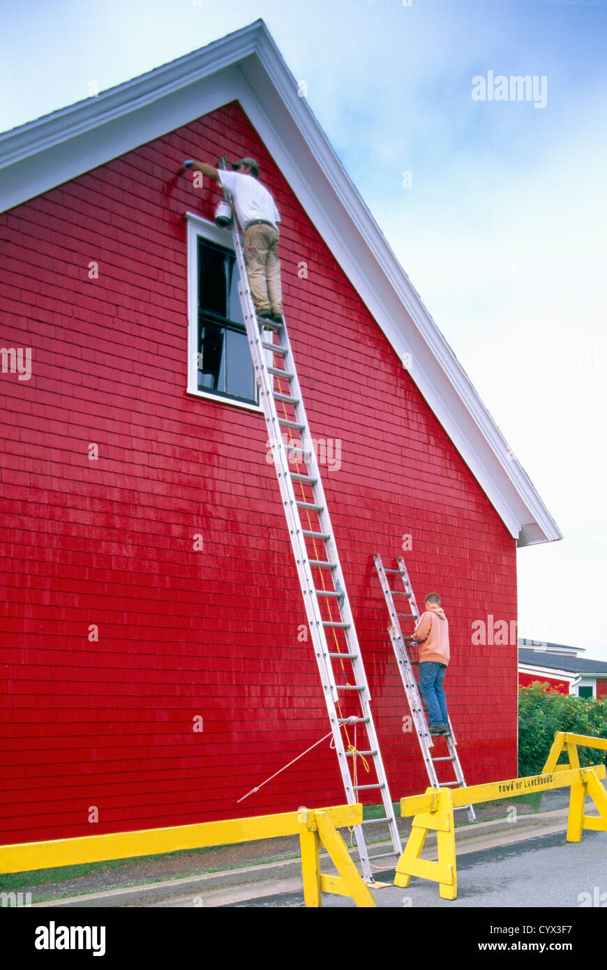 Pittori in piedi su scale, pittura luminosa vernice rossa sulla parete esterna schierandosi in legno di un edificio adibito a magazzino Foto Stock