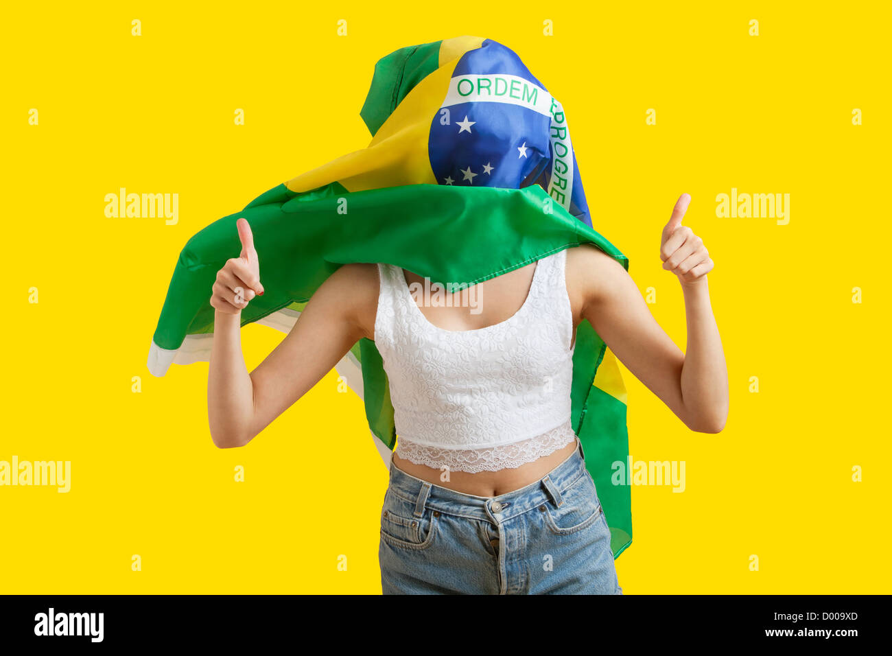 Giovane donna con bandiera brasiliana sulla faccia gesticolando pollice in alto su sfondo giallo Foto Stock
