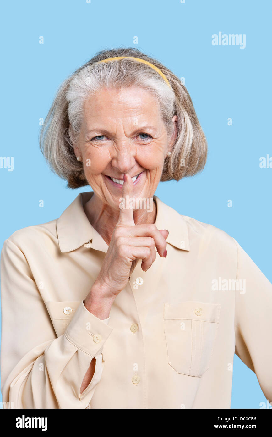 Ritratto di sorridente donna senior in casuals con il dito sulle labbra contro sfondo blu Foto Stock
