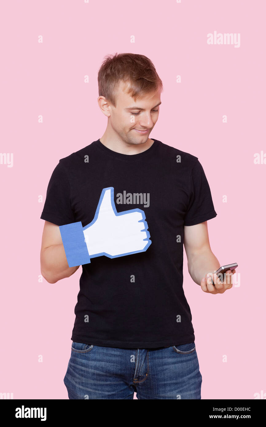 Giovane uomo con un telefono cellulare holding fake like button contro sfondo rosa Foto Stock