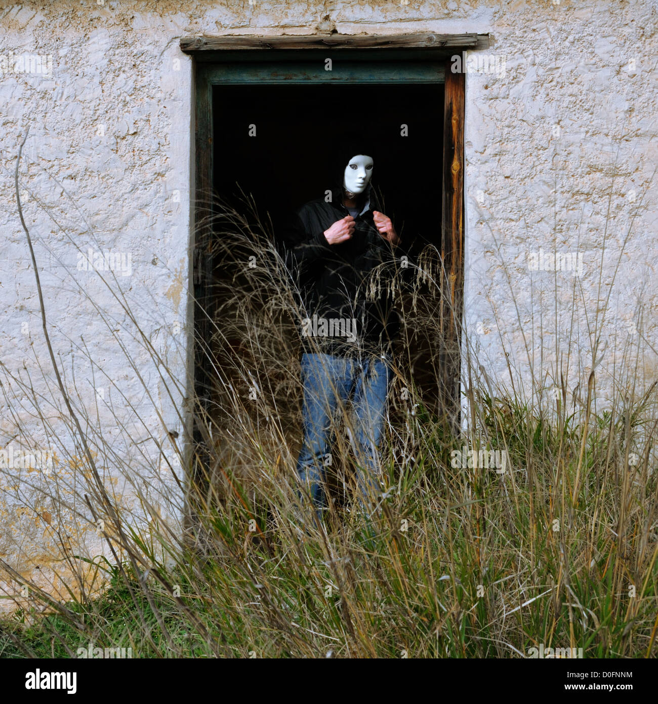 Uomo con maschera bianca dalla rottura di porta della casa abbandonata e ricoperta di piante. Foto Stock