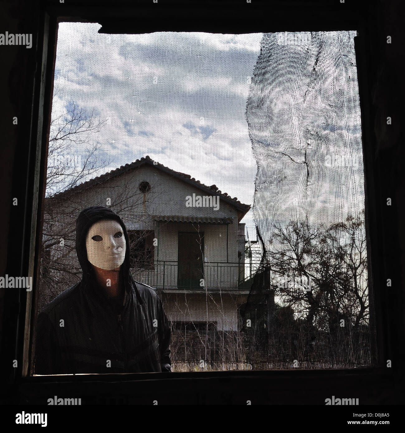 La figura con cappuccio filettato dietro la finestra di casa abbandonati. Masked maniac guardando a voi il viewer! Foto Stock