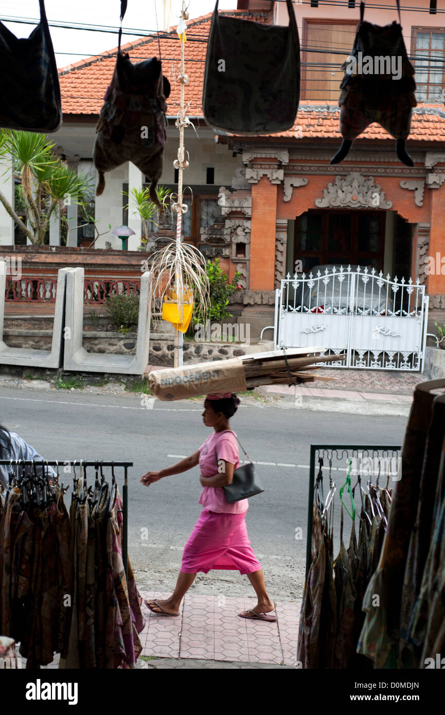 Una donna balinese ,passando da un negozio di vestiti , con un mazzetto di boschi sulla sua testa . Foto Stock