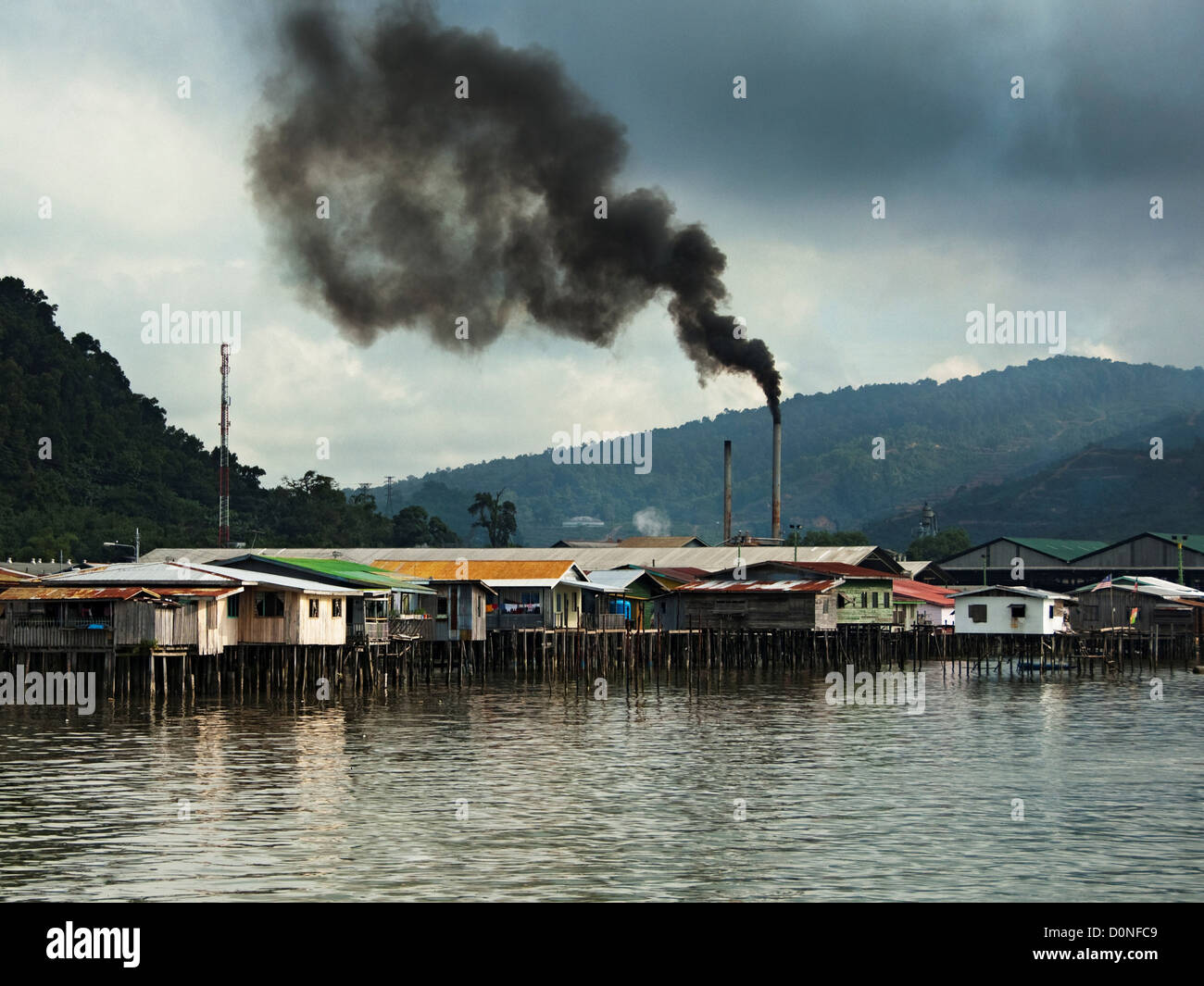 Pennacchi di fumo nero dal camino di fabbrica accanto a un villaggio di acqua, Sandakan, Sabah, Malaysia. Foto Stock