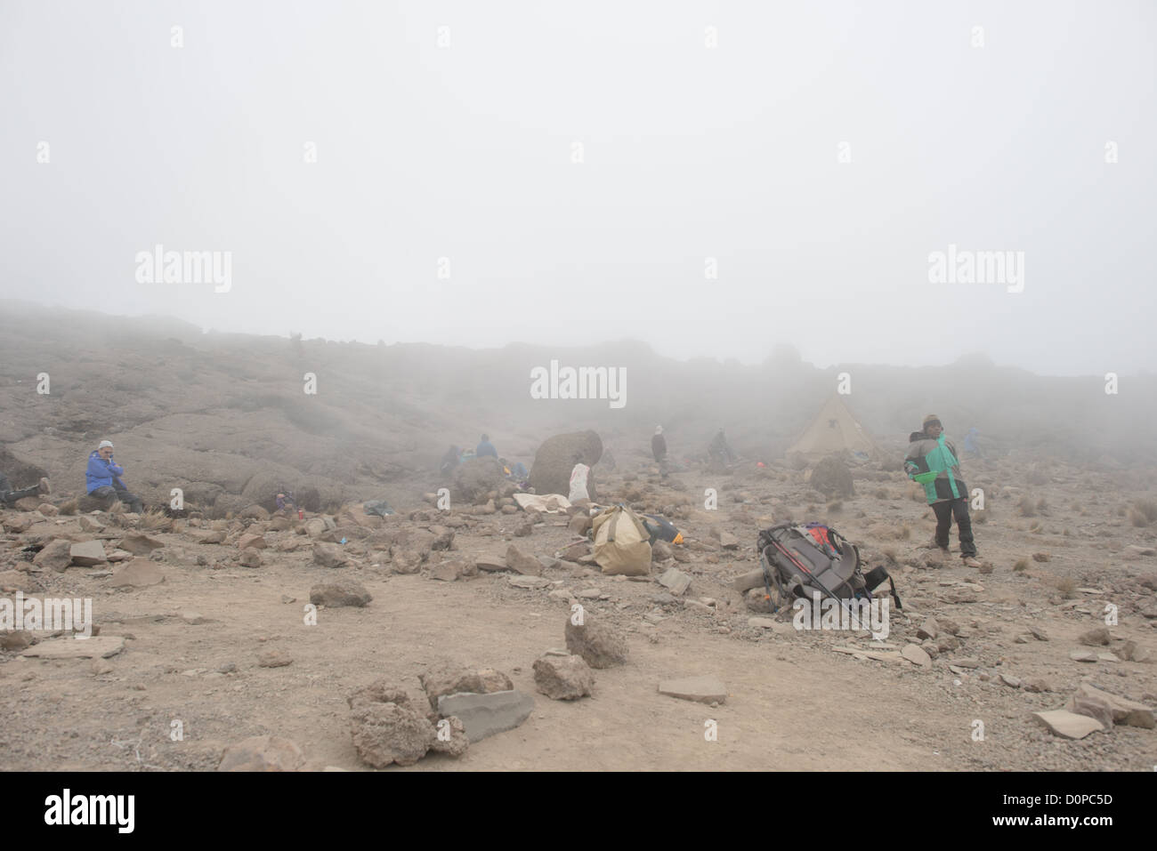 Scalatori del Monte Kilimangiaro Campo Millennium Tanzania | MOUNT KILIMANJARO, Tanzania — Scalatori e portatori si riposano tra l’attrezzatura sparsa nella fitta nebbia vicino al Campo Millennium. Il terreno arido e roccioso segna la transizione nella zona desertica alpina della montagna, dove gli sbalzi estremi di temperatura impediscono la crescita della maggior parte della vegetazione. Questa fitta copertura nuvolosa spesso ricopre i pendii alti quando i venti alisei carichi di umidità dall’Oceano Indiano sono spinti verso l’alto dal massiccio stratovulcano. Foto Stock