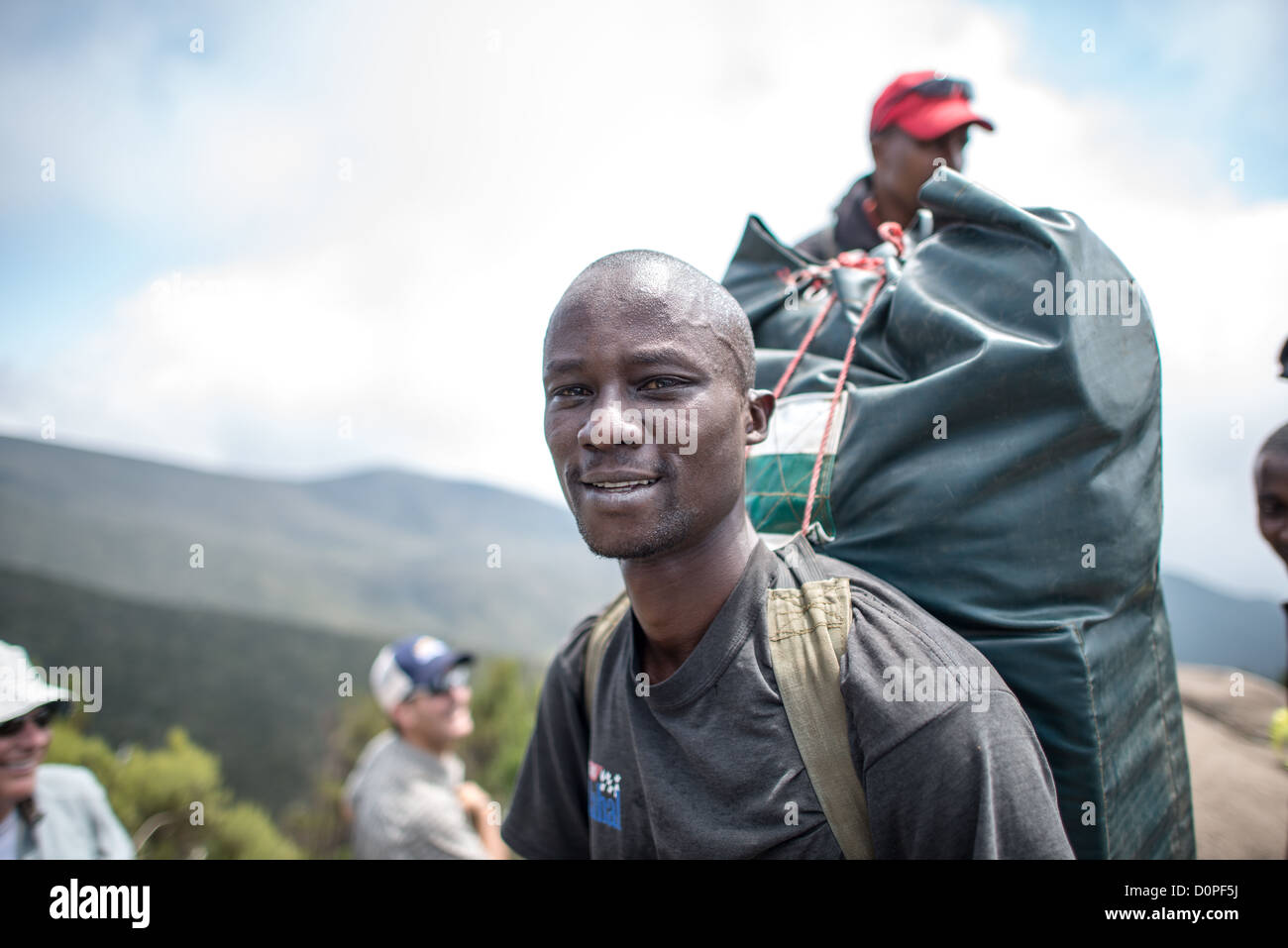 Mount Kilimanjaro Porter Lemosho Trail Tanzania | MOUNT KILIMANJARO, Tanzania — Un portatore trasporta un grande borsone lungo il sentiero Lemosho nella zona di brughiera del Monte Kilimanjaro. Le autorità del Parco Nazionale del Kilimanjaro limitano rigorosamente i carichi individuali a 44 libbre (20 kg) per proteggere i lavoratori dalle lesioni durante l'ascesa di più giorni. La vegetazione circostante a questa quota di 10.000 piedi (3.050 m) segna la transizione ecologica della montagna dalle fitte foreste pluviali basse agli arbusti resistenti della brughiera alpina. Foto Stock