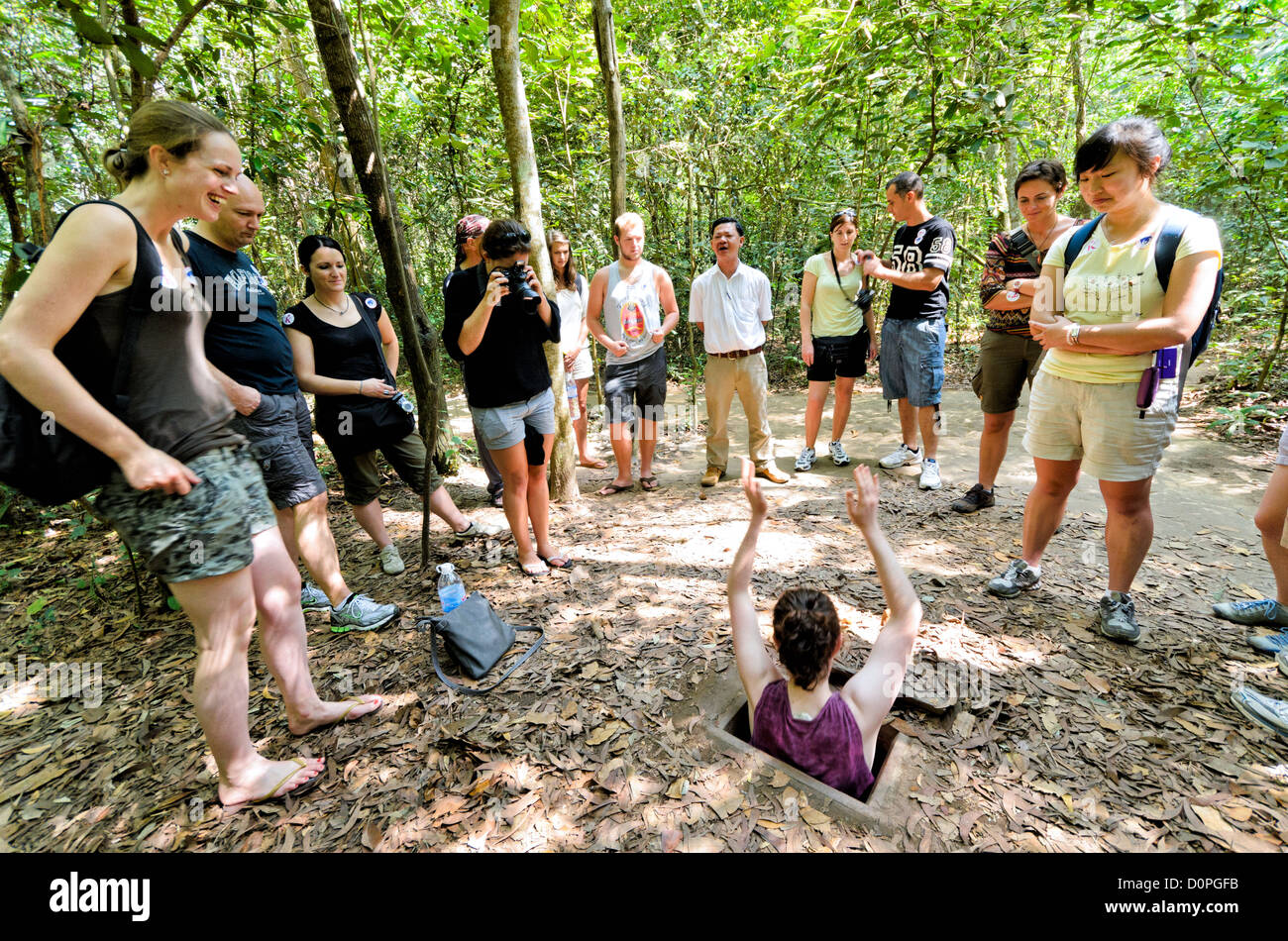 Tunnel di Cu chi Turistico entrando nel tunnel entrata ho chi Minh City Vietnam // CITTÀ DI HO CHI MINH, Vietnam - i turisti tentano di entrare in uno degli ingressi del tunnel preservati nel sito storico di Cu chi Tunnels. L'ingresso mimetico dimostra l'ingegnosità del progetto e della costruzione della rete di tunnel originale. Questi punti di accesso conservati rappresentano parte dell'ampio complesso sotterraneo che si estendeva per oltre 120 chilometri durante la guerra del Vietnam. I tunnel di Cu chi fungevano da rete sotterranea cruciale per le forze vietnamite, fornendo riparo, rotte di rifornimento e vantaggi strategici. Foto Stock