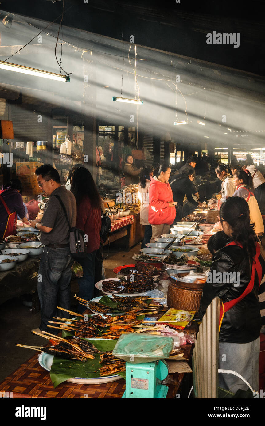 PHONSAVAN, Laos — PHONSAVAN, Laos - la luce del sole del mattino cattura il fumo dalle stufe come venditori al mercato del mattino a Phonsavan nel nord-est del Laos vendere un'ampia gamma di cibo appena cucinato e cucina locale, tra cui passero alla griglia, maiale, pollo, insetti, ratti di bambù, e cane. La popolazione della regione è prevalentemente di etnia Hmong. Foto Stock