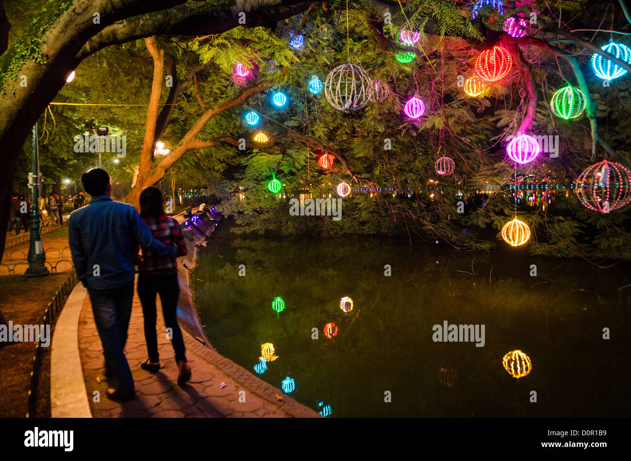 Sentiero illuminato del lago Hoan Kiem Hanoi Vietnam // HANOI, Vietnam - luci colorate sospese dagli alberi illuminano il sentiero costiero intorno al lago Hoan Kiem nel centro di Hanoi. Una coppia cammina lungo il lungolago sotto la tettoia illuminata. Lo storico lago, situato nel cuore del centro storico di Hanoi, è un popolare luogo di ritrovo serale. Foto Stock