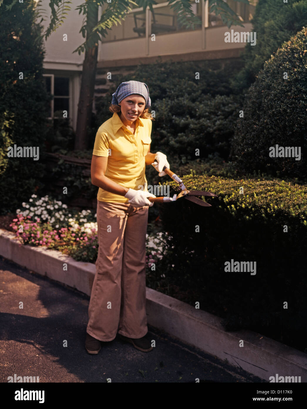 Anni Settanta DONNA CON GIARDINO CLIPPERS TRIMMING siepe di bosso indossando BELLBOTTOMS Foto Stock