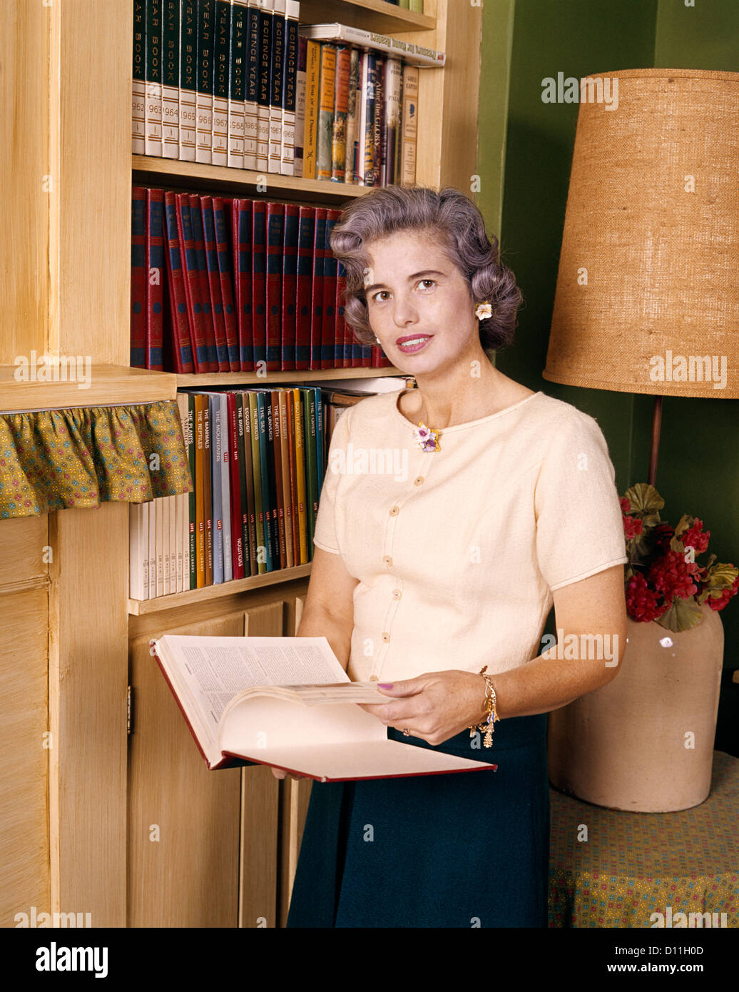 Anni sessanta donna di mezza età di lettura aperta di contenimento ENCYCLOPEDIA PRENOTA sorridente in piedi dalla libreria di casa guardando la fotocamera Foto Stock
