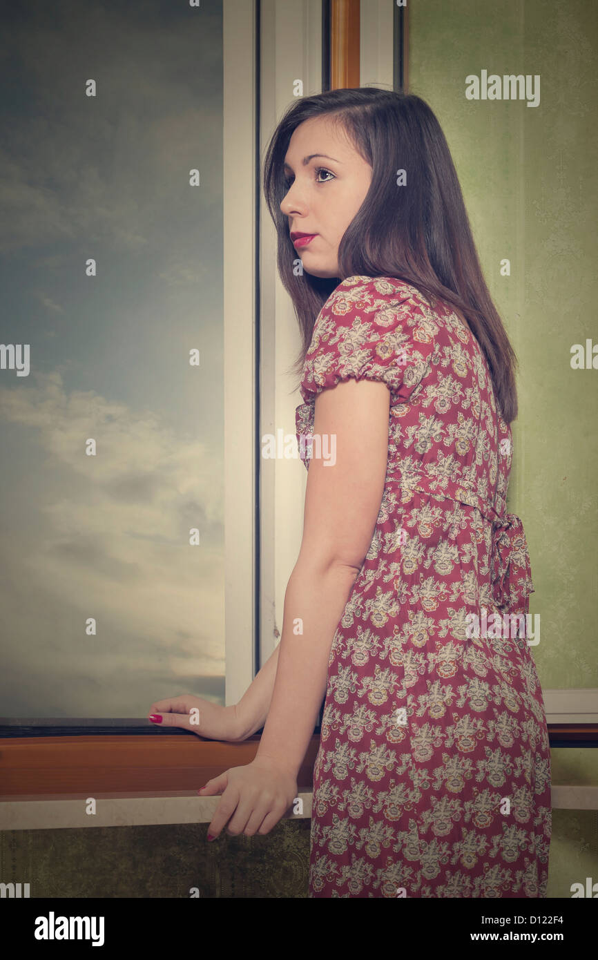 Una ragazza in un abito floreale è in piedi accanto a una finestra aperta Foto Stock