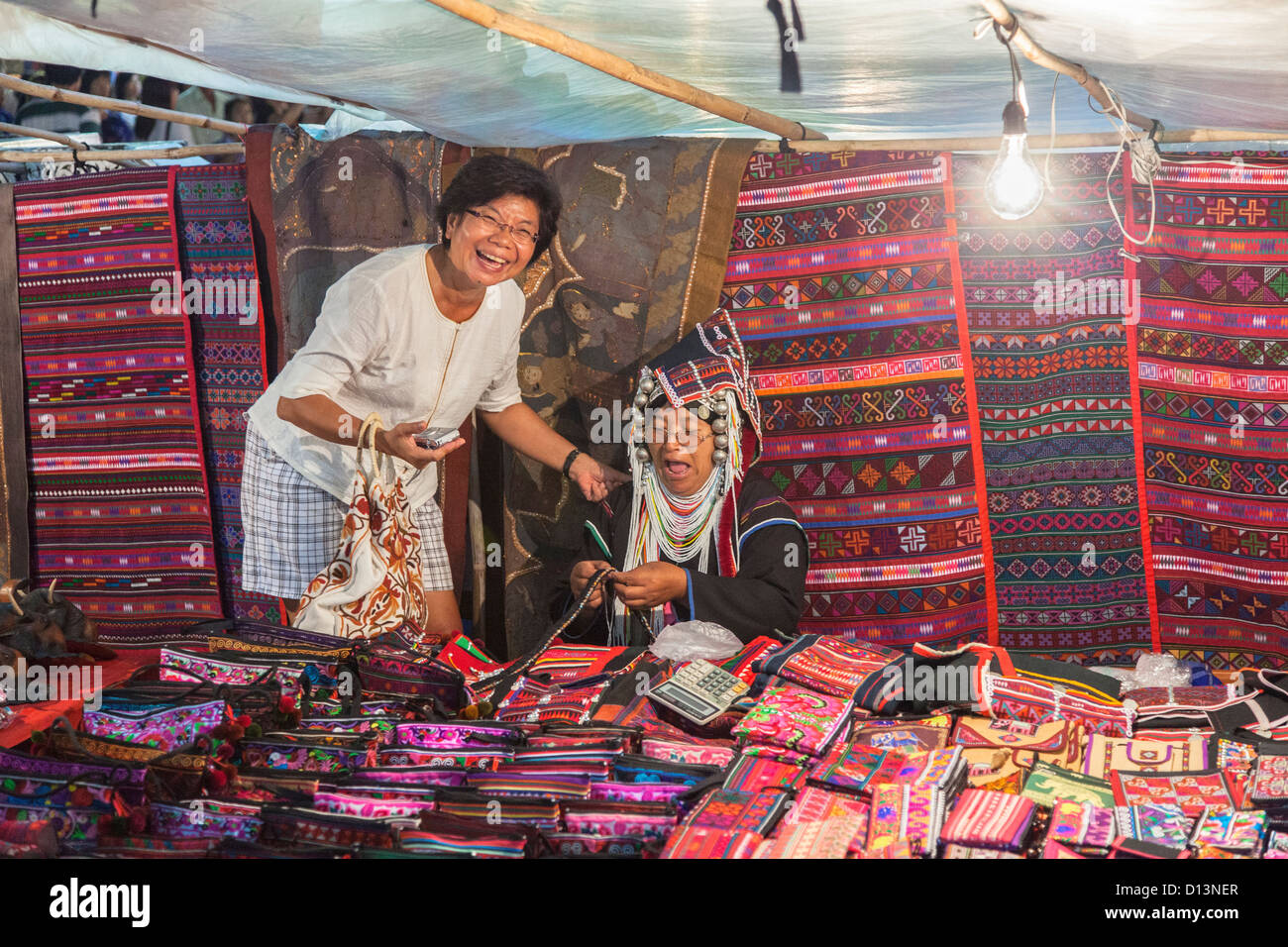 Stallo arazzi di vendita al mercato notturno di Chiang Rai, la Thailandia del Nord, due donne locali ridendo e godendo di uno scherzo Foto Stock