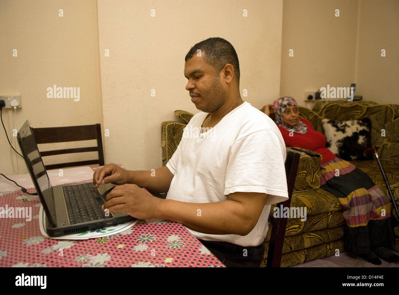 Il marito che si prende cura di sua moglie disabili a casa (seduto, amputazione della gamba) utilizza il computer portatile mentre mia moglie guarda la televisione Foto Stock