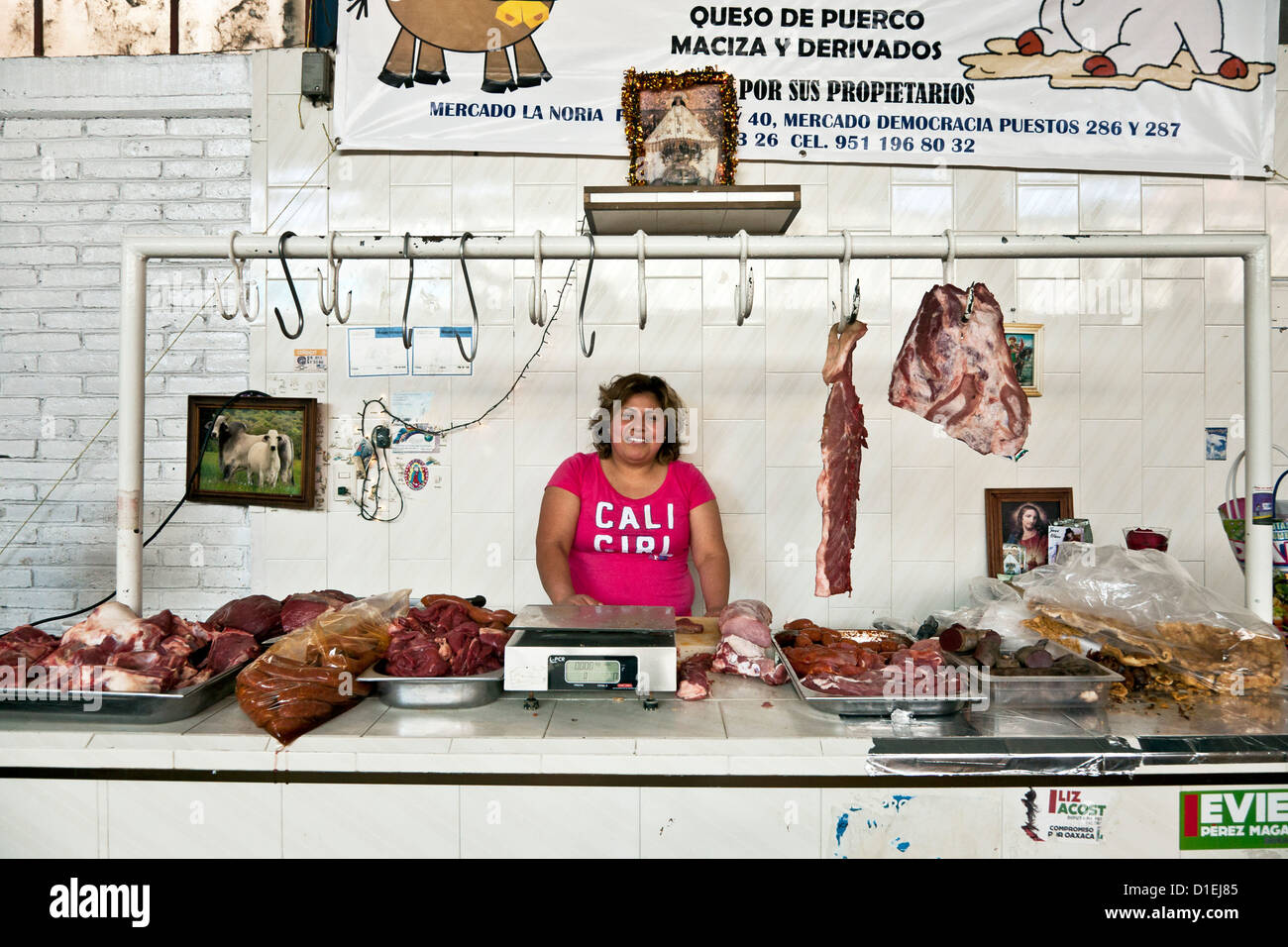 Paffuto donna sorridente macellaio dietro il bancone con vaschette di carne & haunches appesi al soffitto rack sotto il piccolo santuario Oaxaca Foto Stock