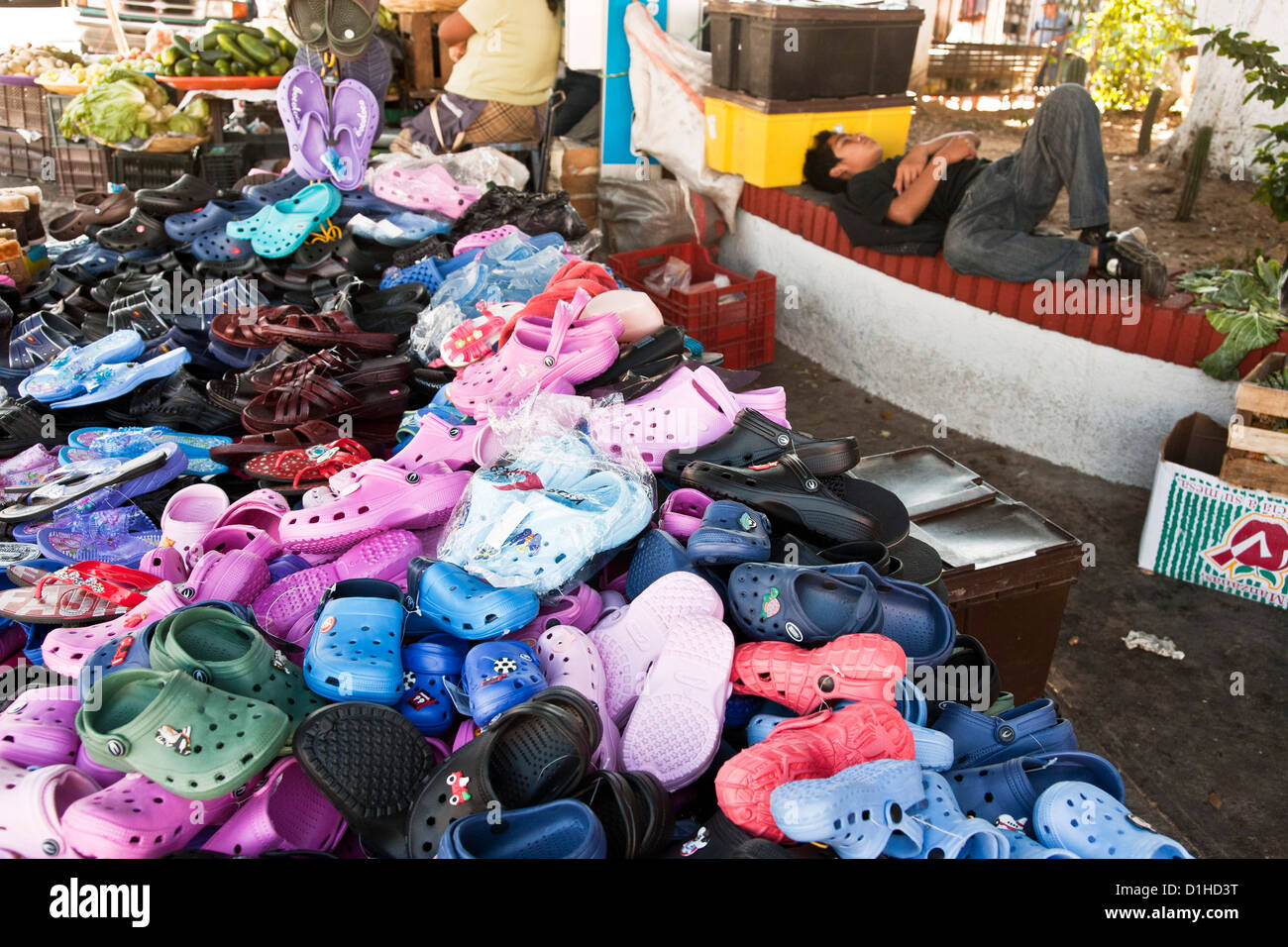 Un grande e luminoso pila di croc tipo le scarpe per la vendita con snoozing Mexican teen venditore prendendo una pausa mercado La Merced mercato Oaxaca Foto Stock