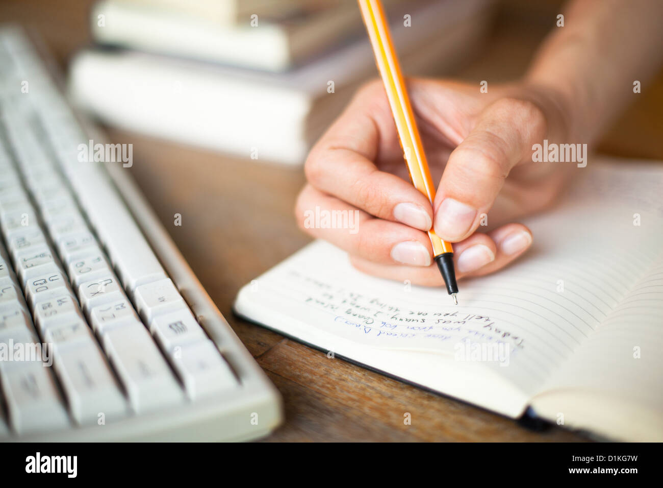 Foto di mani scrive una penna in un notebook, la tastiera del computer e una pila di libri in background Foto Stock