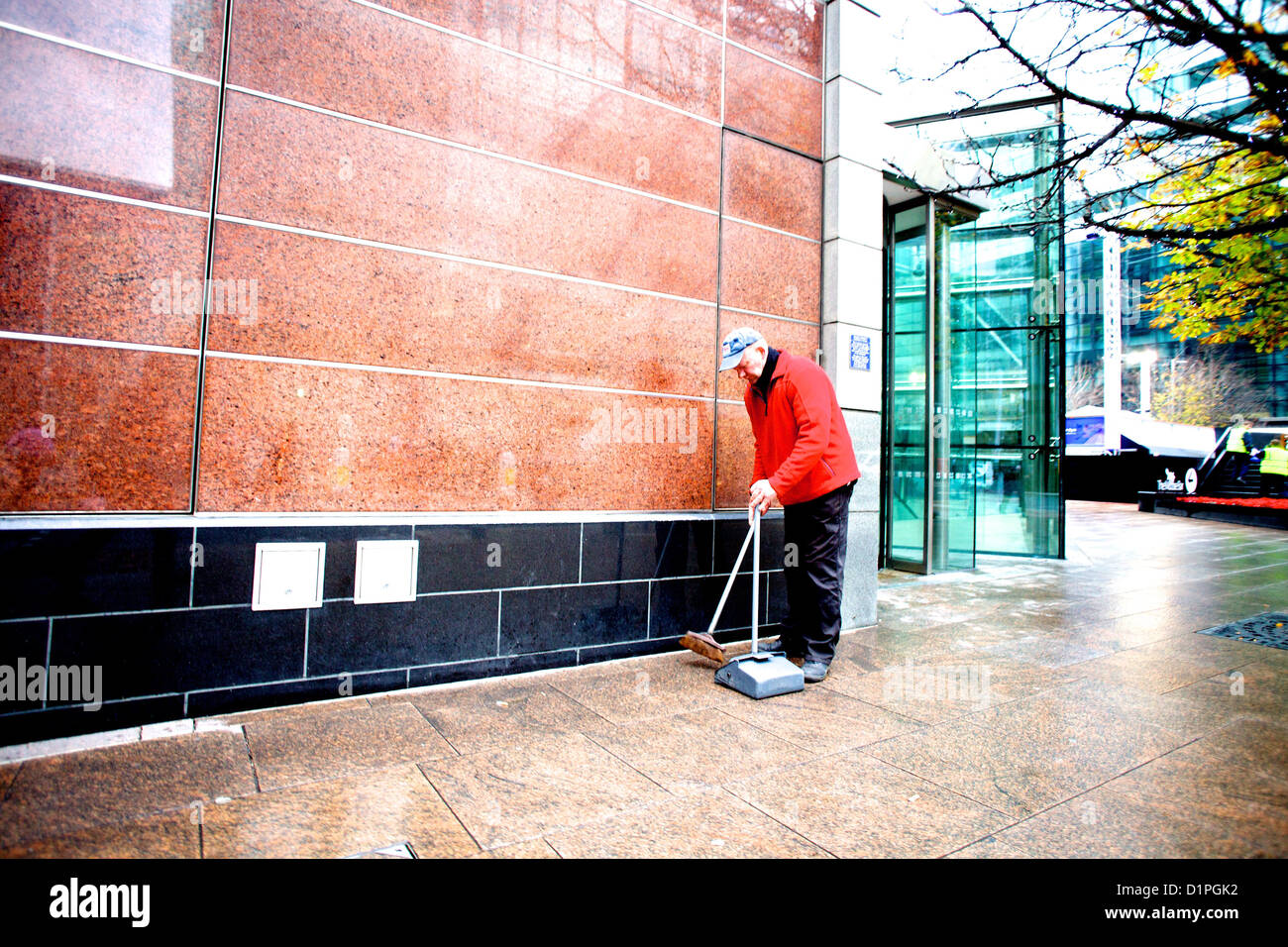 Uomo anziano in giacca rossa spazzando dei rifiuti nel contenitore attorno ad un blocco di uffici di Canary Wharf, Londra. Foto Stock