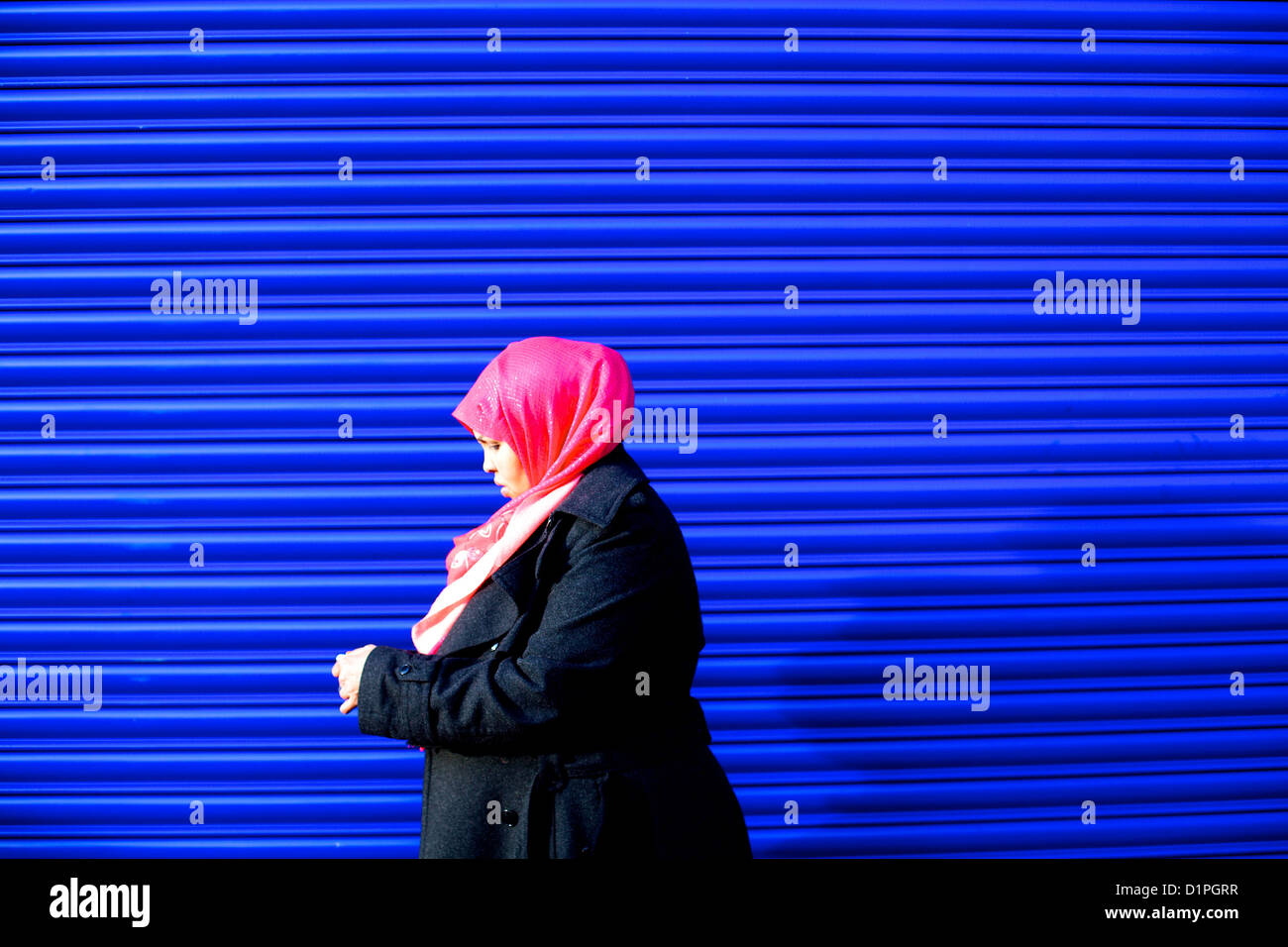 Una donna musulmana guardando il suo telefono cellulare con velo di colore rosa passato a piedi un brillante, blu royal otturatore Foto Stock