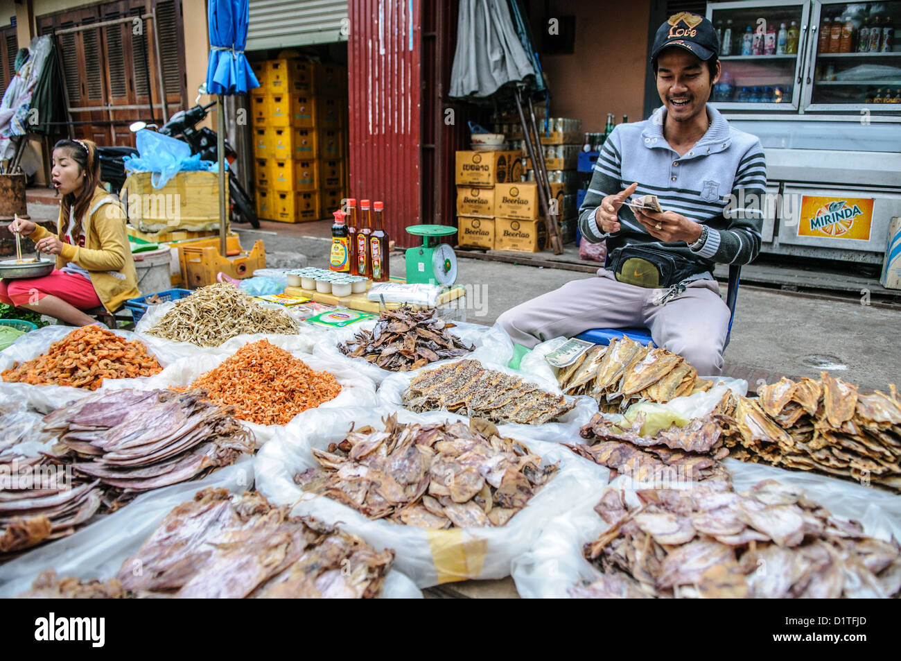 Pesce secco al mercato mattutino di Luang Prabang Laos // LUANG PRABANG, Laos - Un venditore propone pesce secco e salato, tra cui calamari, gamberi e pesce al mercato mattutino. Questi prodotti conservati servono come ingredienti essenziali nella cucina tradizionale del Laos, fornendo fonti proteiche che possono essere conservate senza refrigerazione nel clima tropicale. Le tecniche di conservazione dell’essiccazione e della salatura consentono lo stoccaggio e il trasporto a lungo termine dei prodotti ittici dalle zone costiere ai mercati interni del Laos. Il mercato mattutino di Luang Prabang funge da centro centrale per il commercio locale, dove Foto Stock
