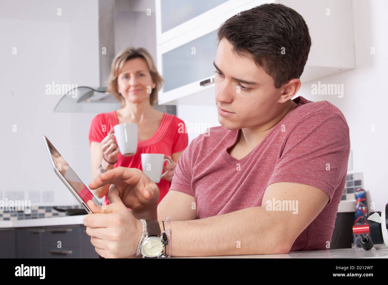 Giovane uomo utilizzando una tavoletta digitale guardando lo schermo o monitor. Situato in una cucina moderna con una donna che mantiene due tazze sorrisi. Foto Stock