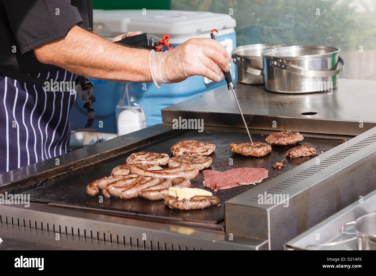 Uomo che indossa guanti di plastica la verifica di un burger per vedere se la cottura completa mentre la cottura di salsicce hamburger bistecca su un cibo piastra di stallo. Inghilterra, Regno Unito Foto Stock
