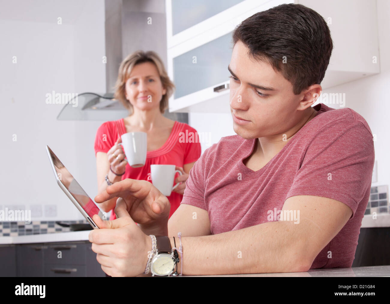 Giovane uomo utilizzando una tavoletta digitale guardando lo schermo o monitor. Situato in una cucina moderna con una donna che mantiene due tazze sorrisi. Foto Stock