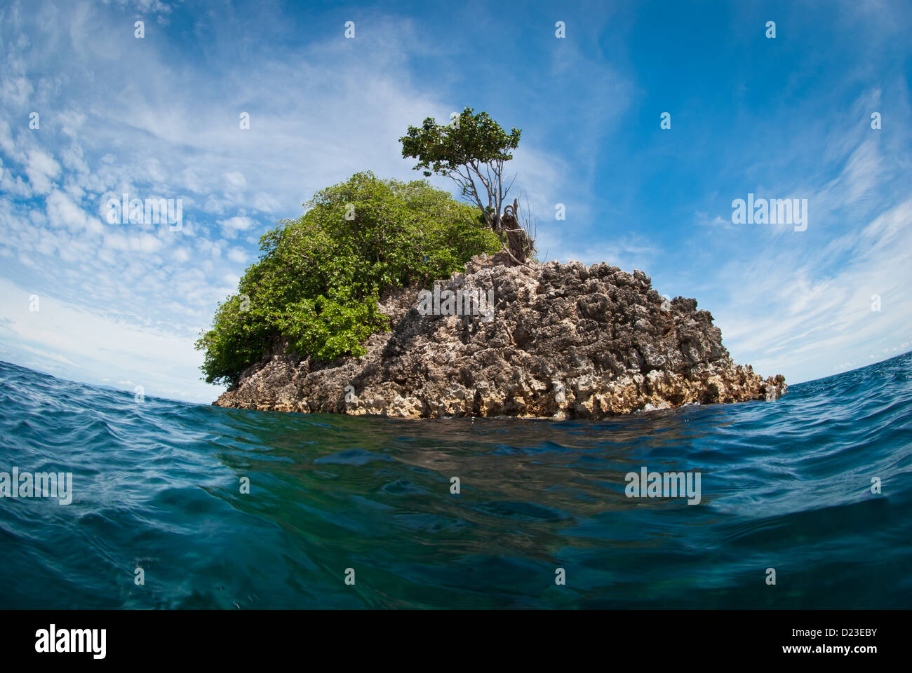 Una piccola isola rocciosa nel mezzo dell'oceano durante una giornata di sole con cielo blu soffici nuvole bianche e calmo mare piatto Foto Stock