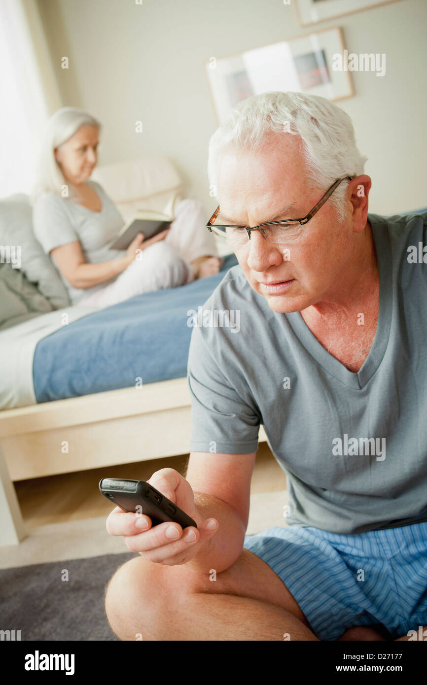 Senior uomo utilizzando il telefono cellulare e il senior donna leggendo un libro sul letto Foto Stock