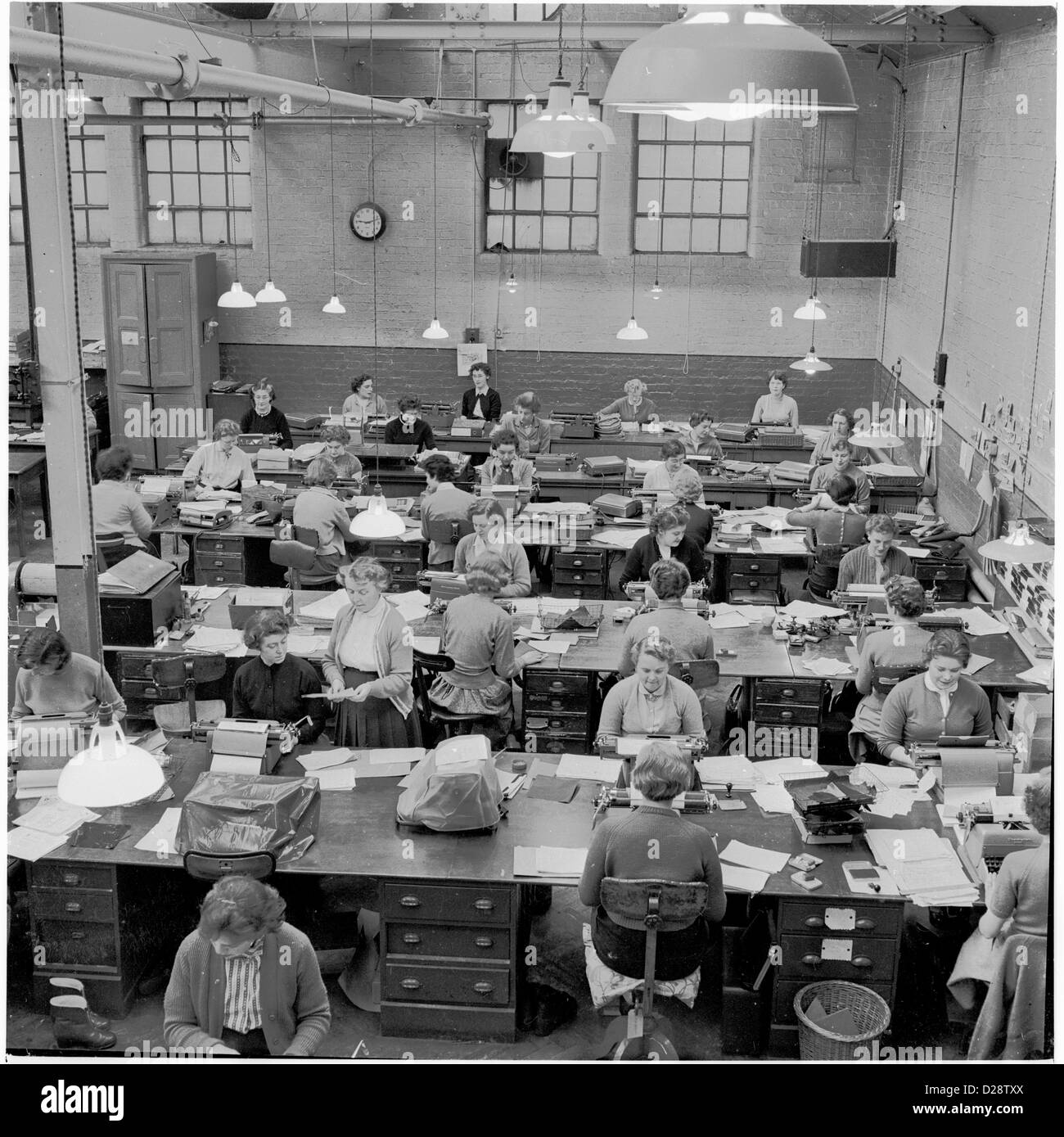 Anni '50, storico. Un pool di digitazione in una fabbrica. Personale di segreteria femminile seduto a grandi scrivanie al lavoro in un piano aperto, area di alta cella di un edificio di fabbrica. Foto Stock