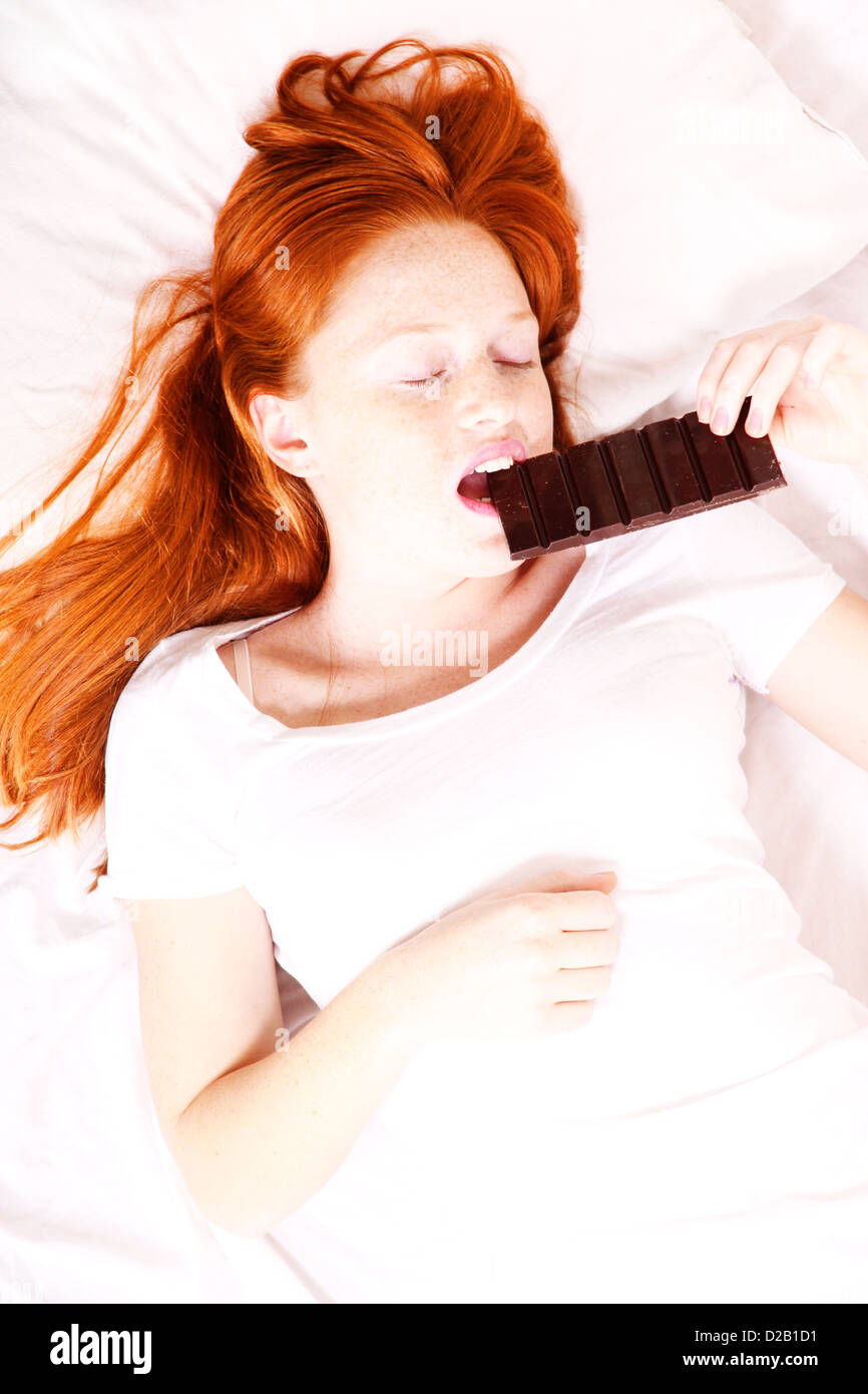 Una giovane donna redhead mangiare il cioccolato a letto. Foto Stock