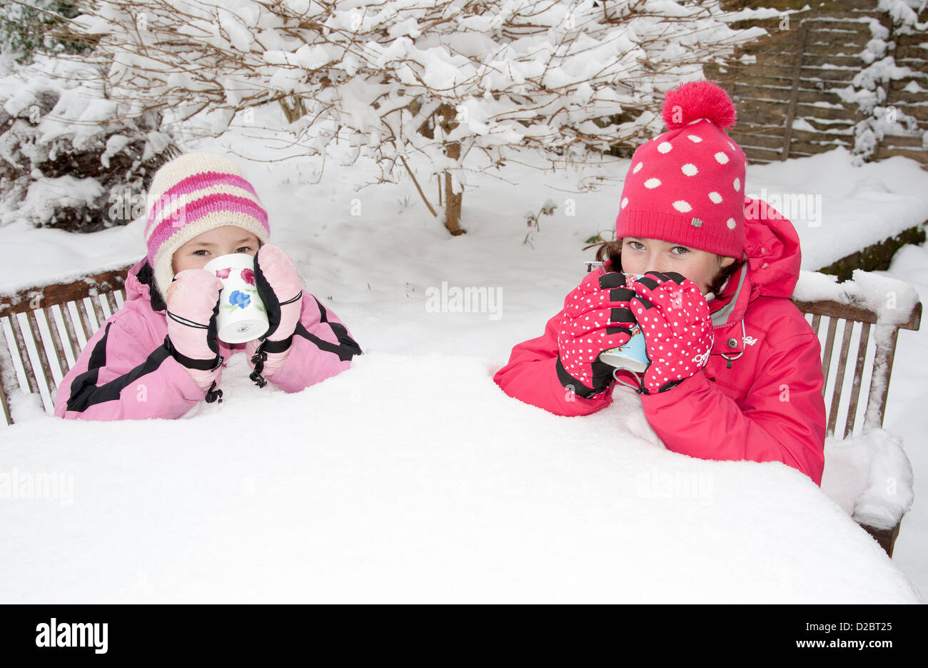 Due giovani ragazze hanno una bevanda calda seduti sulla coperta di neve mobili da giardino durante il periodo invernale nel sud dell'Inghilterra Foto Stock