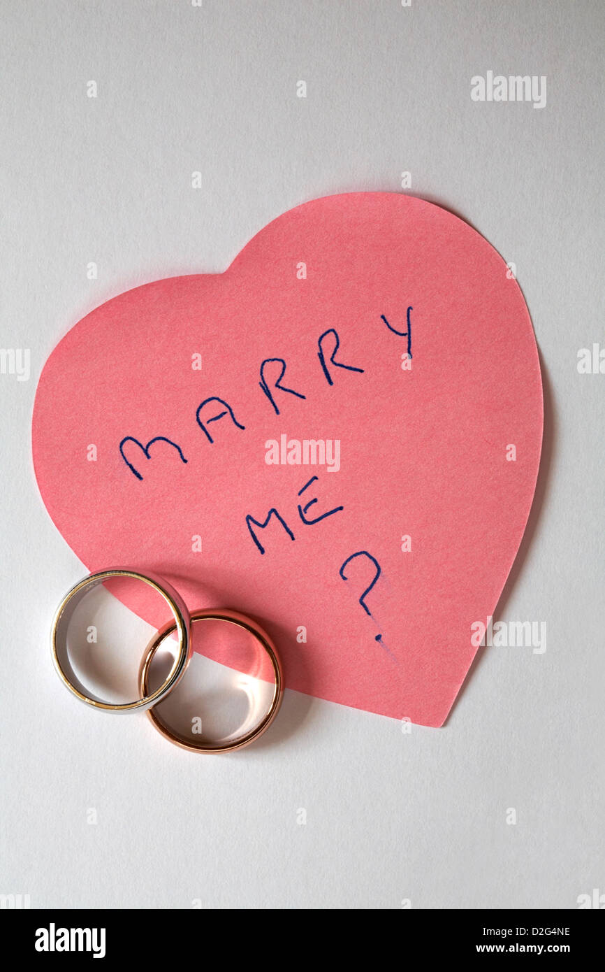 Sposarsi me? Messaggio scritto sul rosa a forma di cuore post it pad con due anelli di nozze - proposta di matrimonio isolati su sfondo bianco Foto Stock