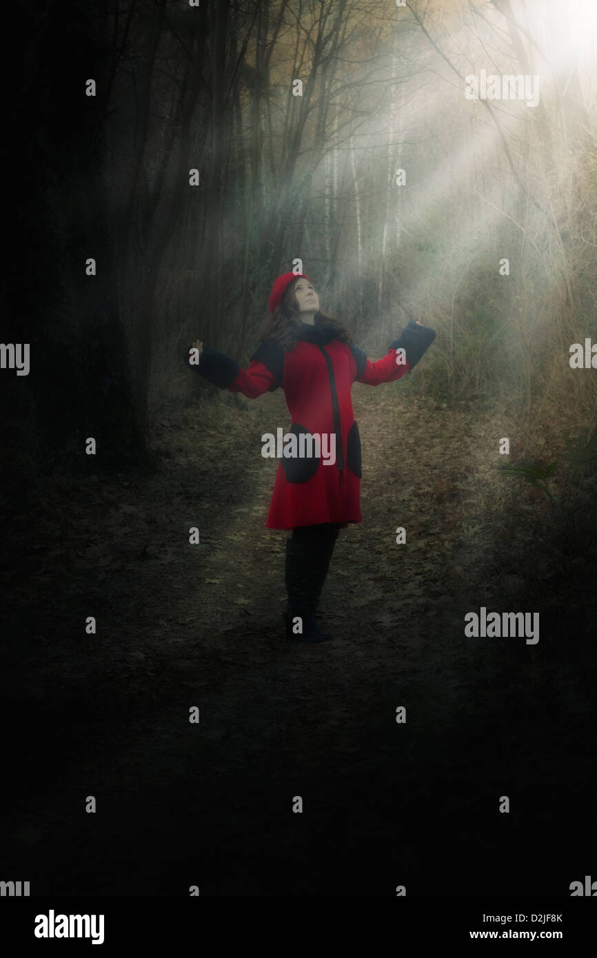 Una donna in un rivestimento di colore rosso è in piedi in una foresta scura in uno spot di luce solare Foto Stock