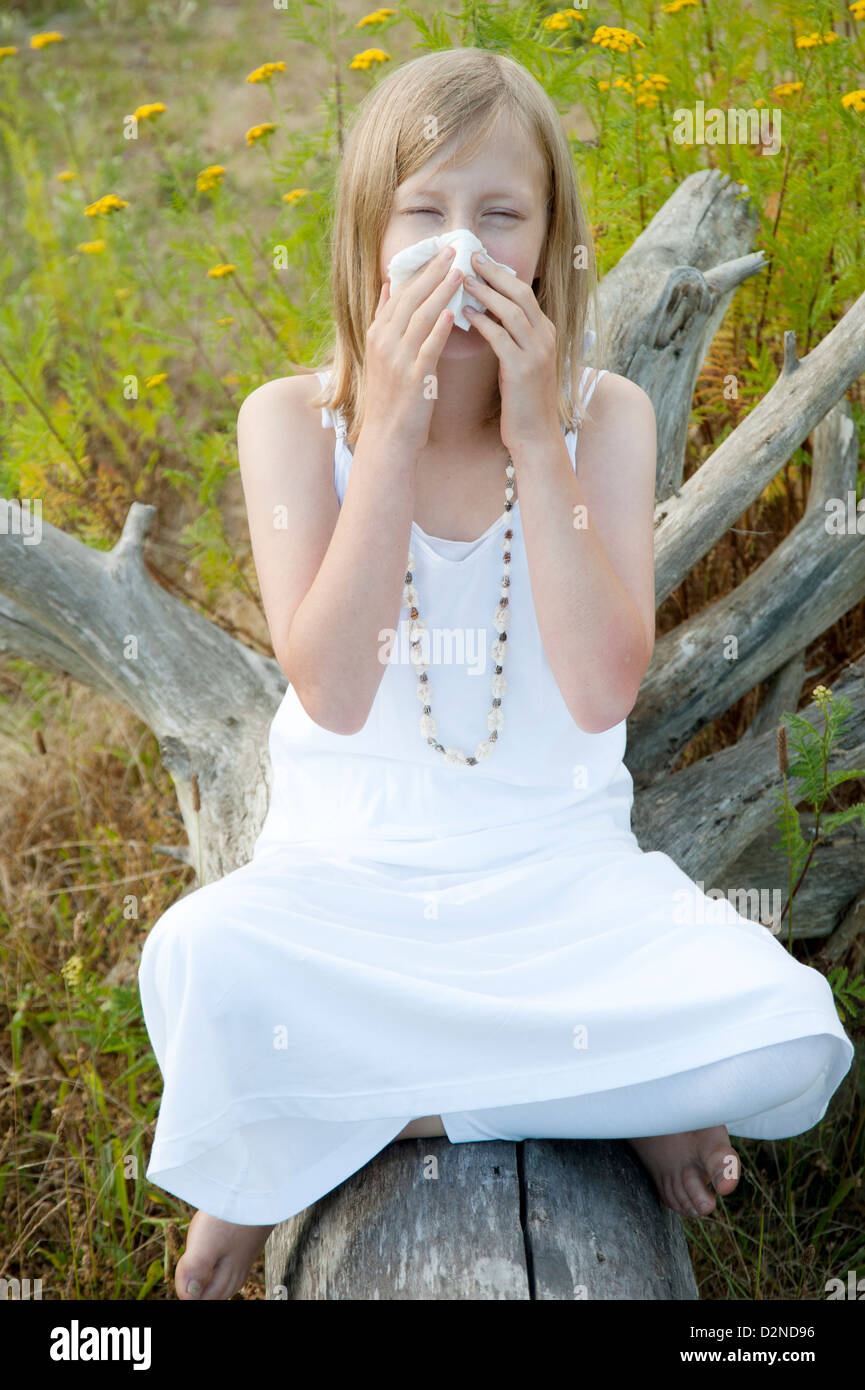 Giovane ragazza seduta su un log in estate circondata da erbacce e soffiare il naso a causa di allergie Foto Stock
