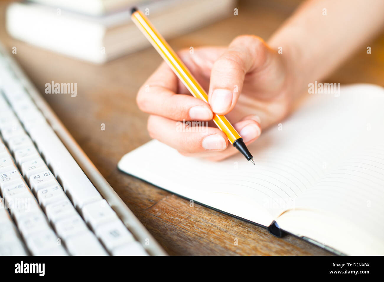 Mani scrive una penna in un notebook, la tastiera del computer e una pila di libri in background Foto Stock