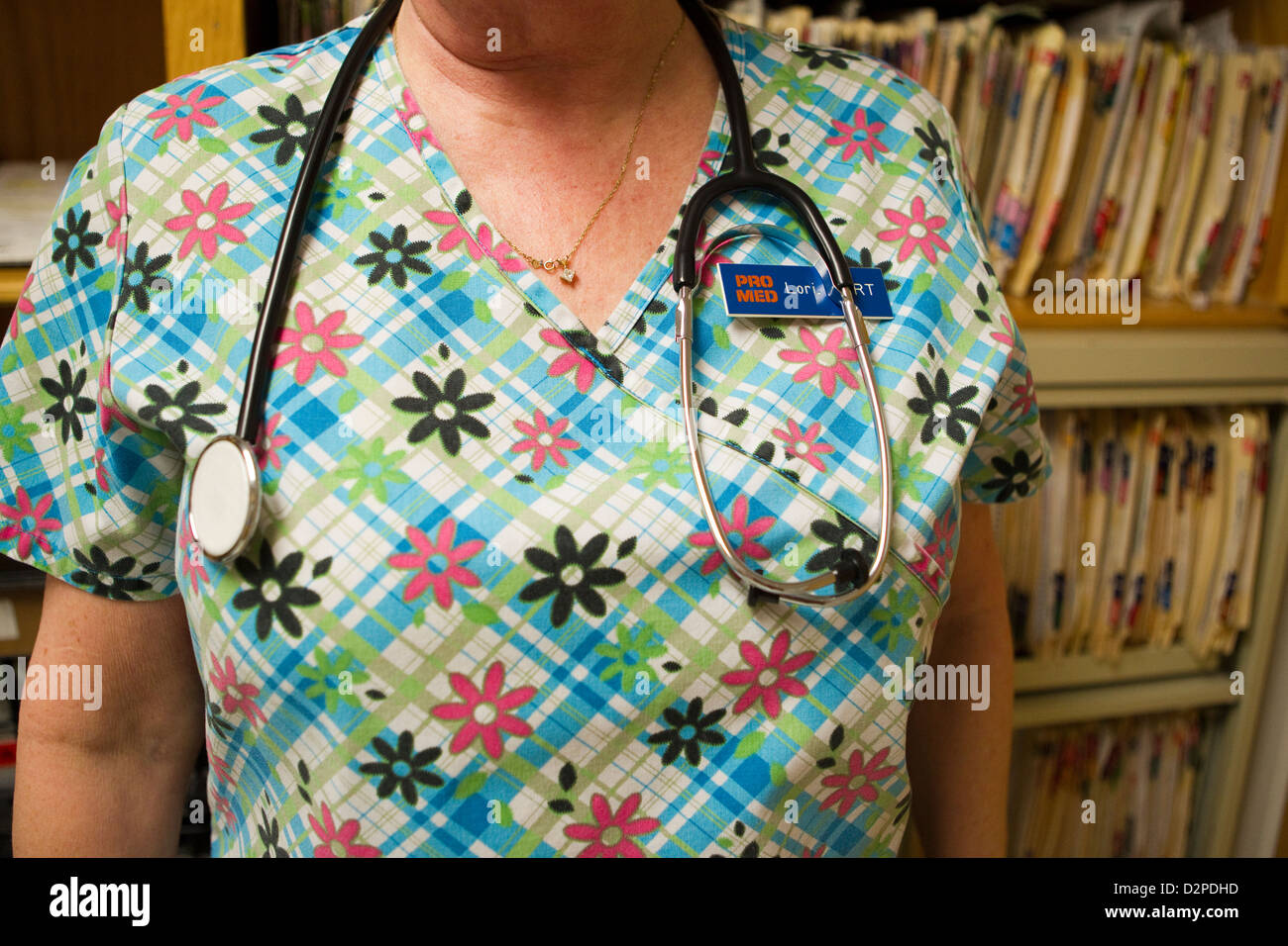 Femmina tecnico medico indossando scrubs e stetoscopio pone nella parte anteriore della parete di cartelle mediche presso una piccola clinica medica Foto Stock