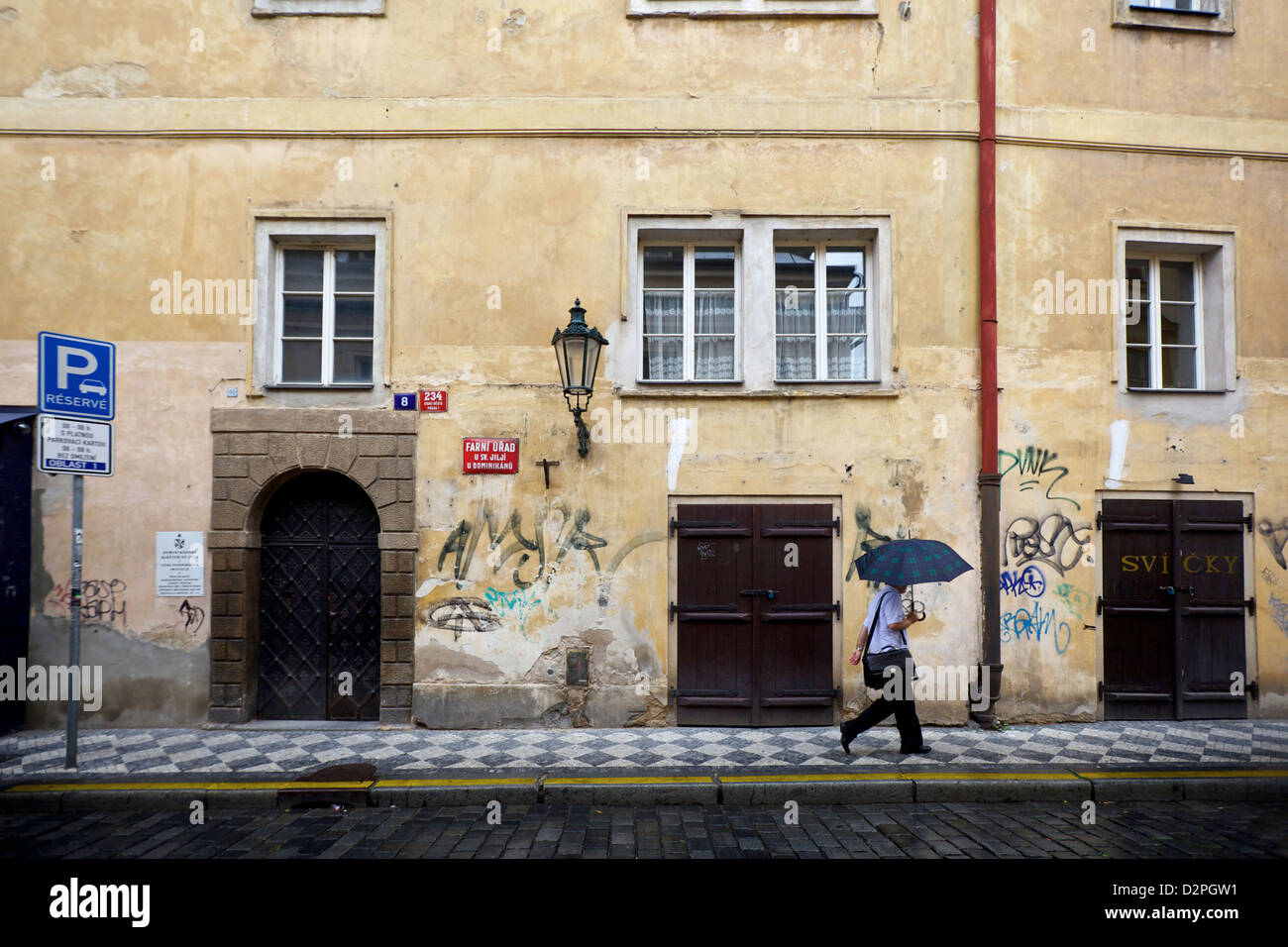 Una figura solitaria con un ombrello cammina lungo una piovosa strada acciottolata a Praga, passando davanti a un edificio storico con pareti coperte di graffiti e vinta Foto Stock