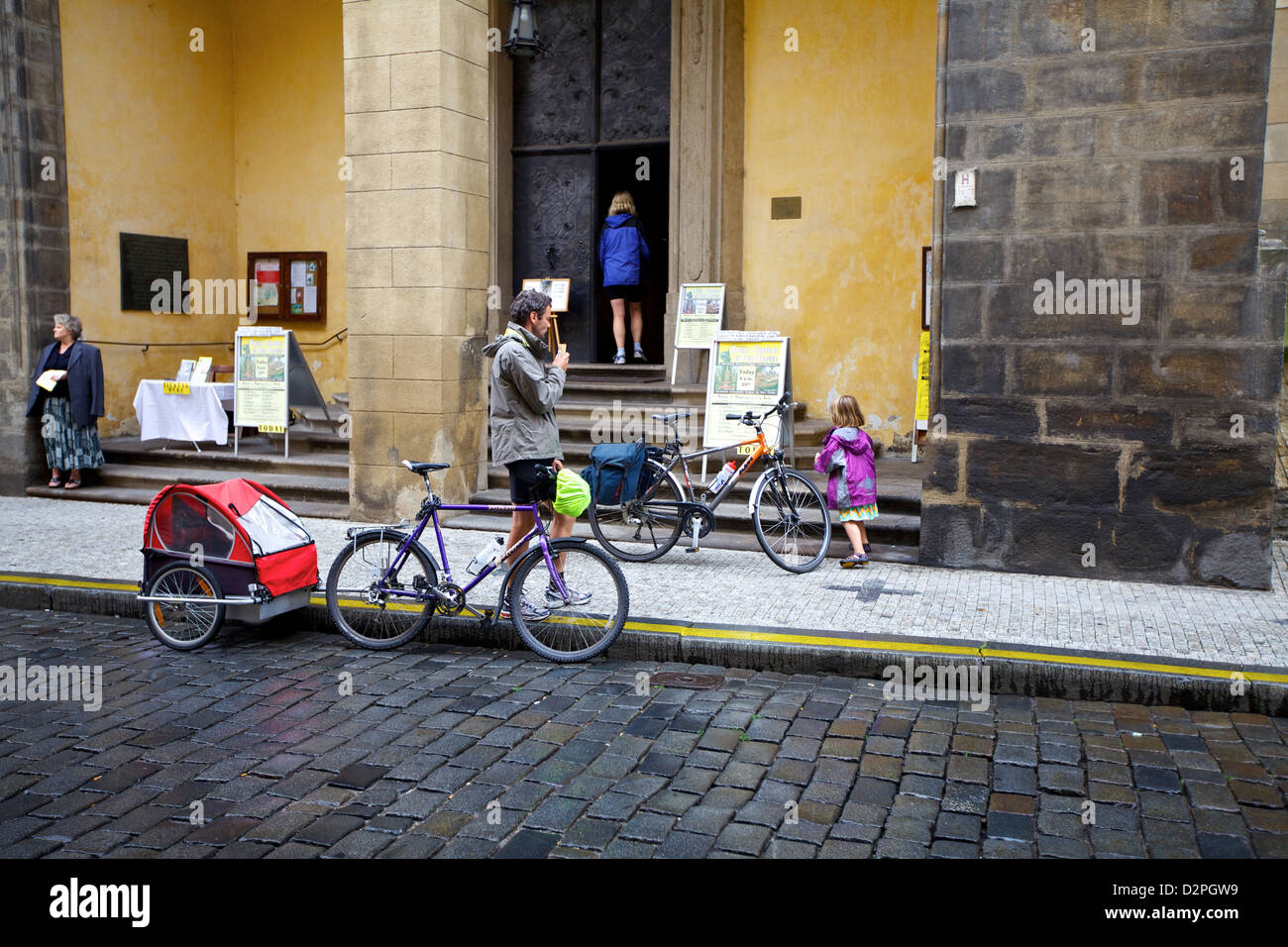 Una famiglia fa una pausa all'esterno di un edificio storico su una strada acciottolata a Praga, con biciclette e un rimorchio per bambini che riflette la miscela di storia della città Foto Stock