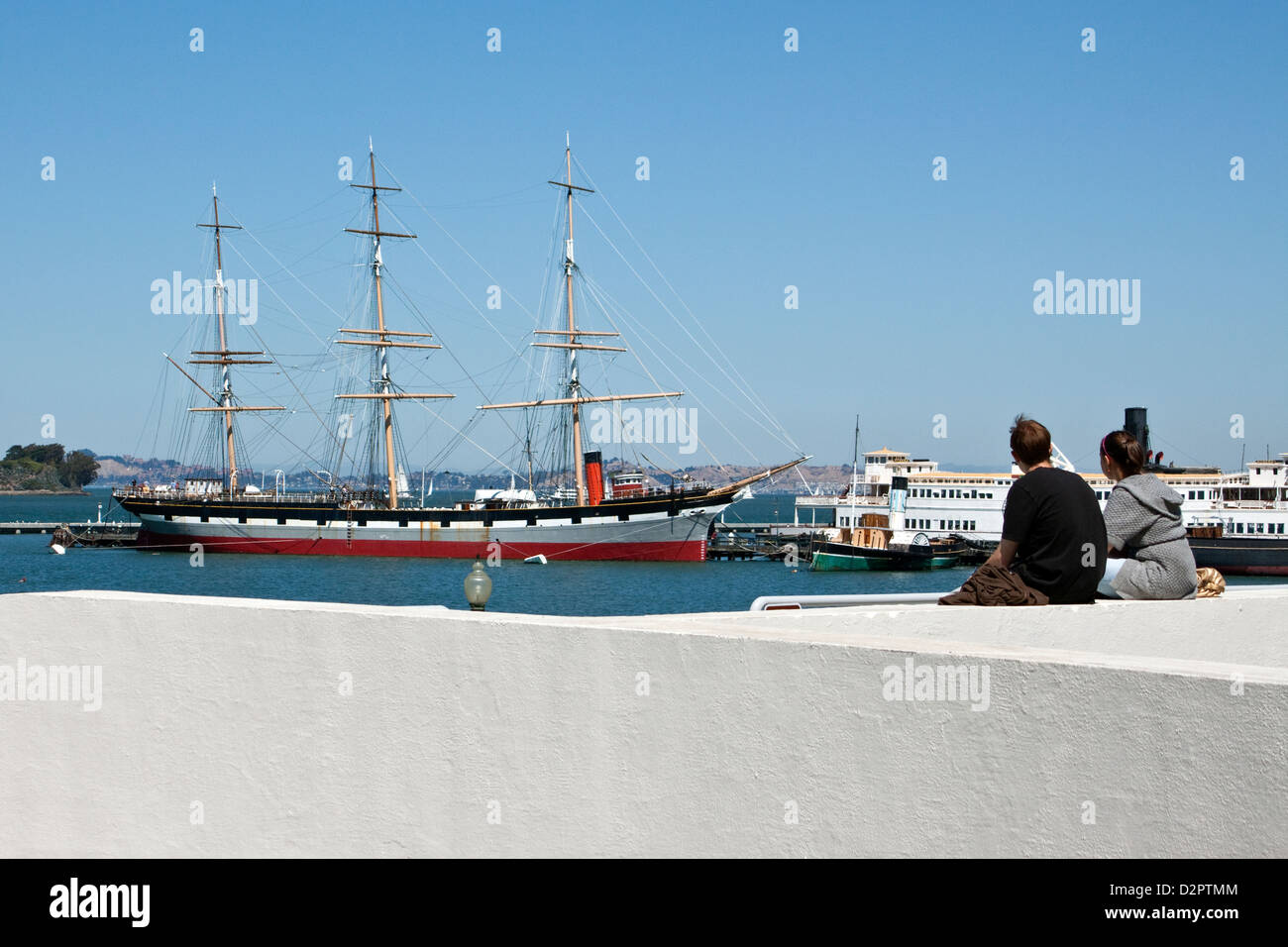 Coppia giovane seduto su una parete del porto guardando verso navi storiche a Hyde Street Pier vicino a Ghirardelli Square a san Francisco, California Foto Stock