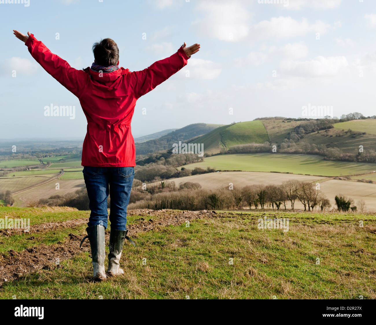 Una donna in un cappotto rosso jeans blu e verde stivali da pioggia sollevando le braccia in un modo celebrativo mentre passeggiate Foto Stock