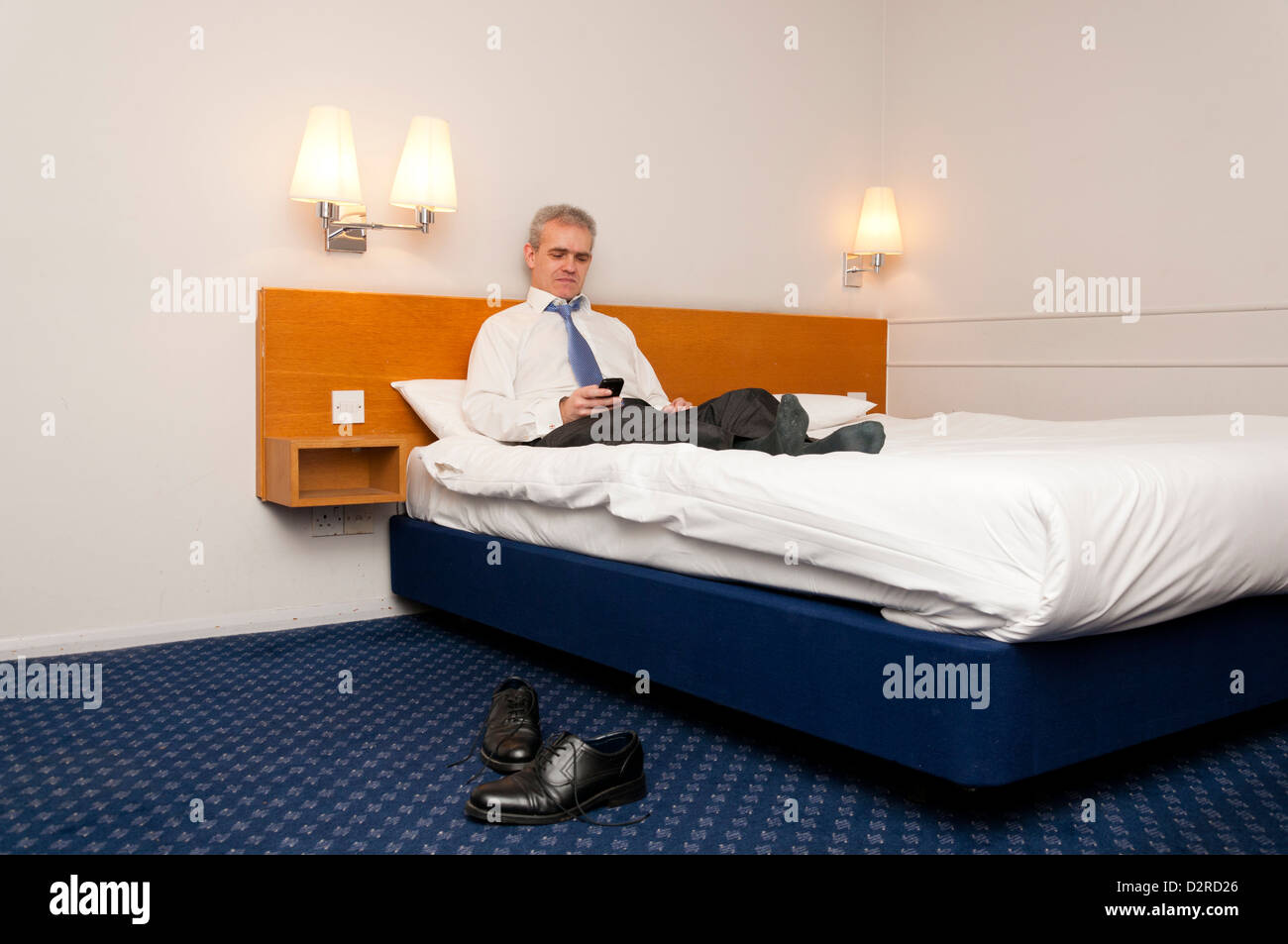Business uomo seduto sul letto hotel controllando il suo telefono per i messaggi. Foto Stock