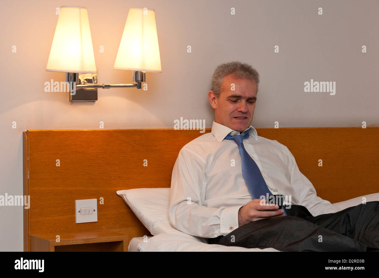 Business uomo seduto sul letto hotel controllando il suo telefono per i messaggi. Foto Stock